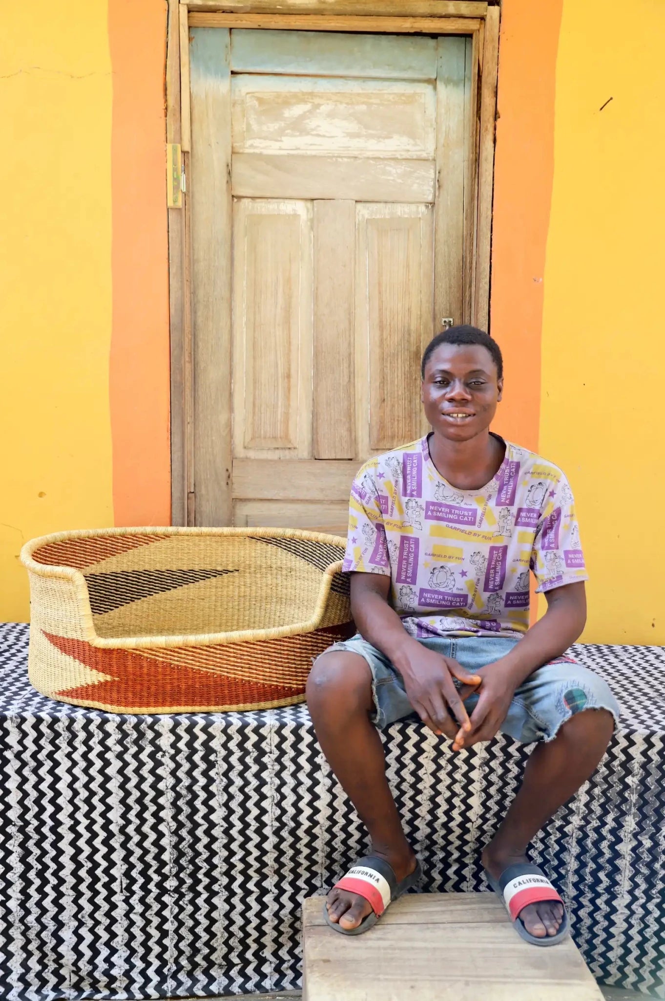  Man  sitting on a bench with a woven basket next to a wooden door and yellow wall.