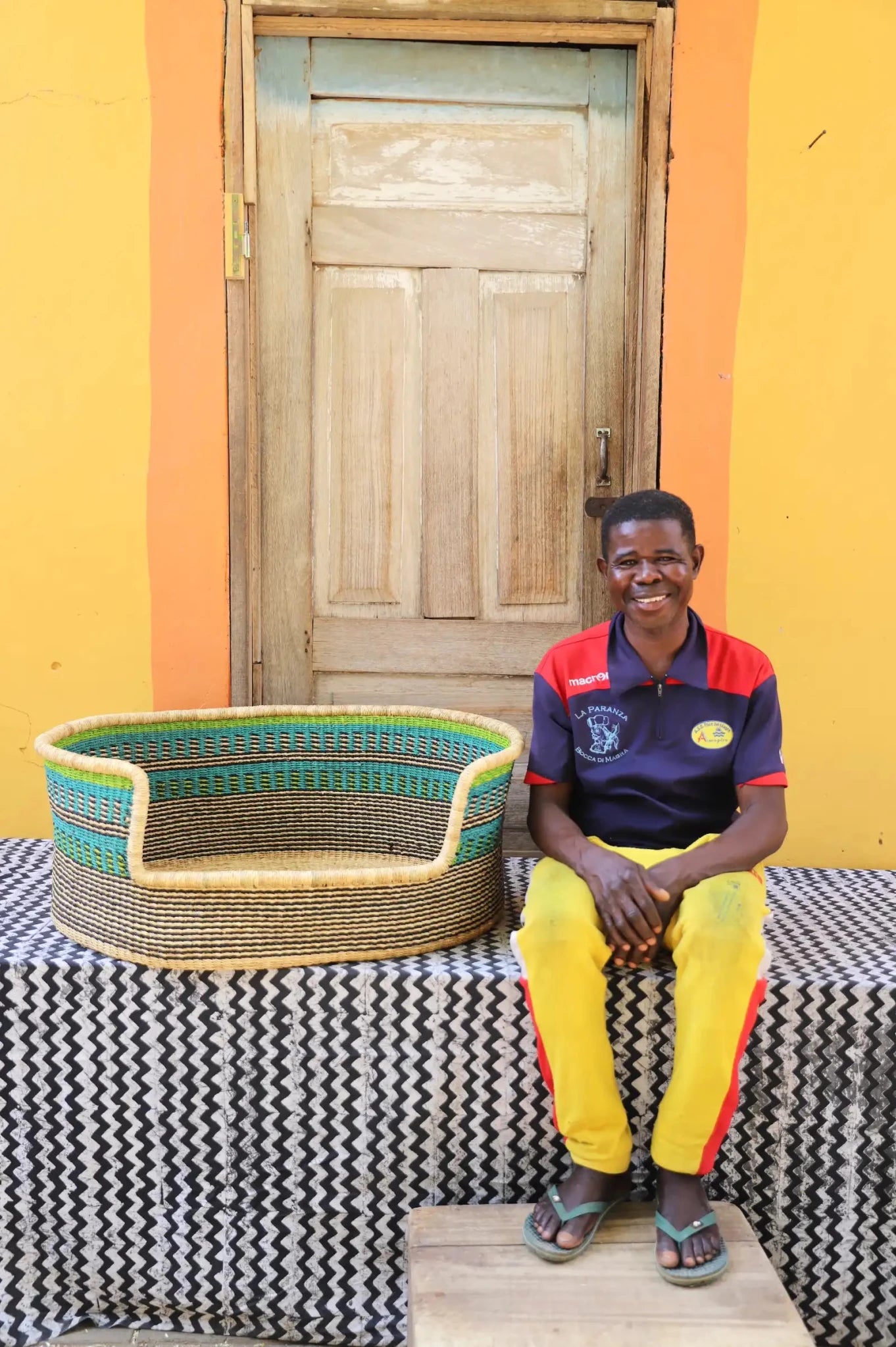 Man  sitting on a bench with a woven basket next to a wooden door and yellow wall.