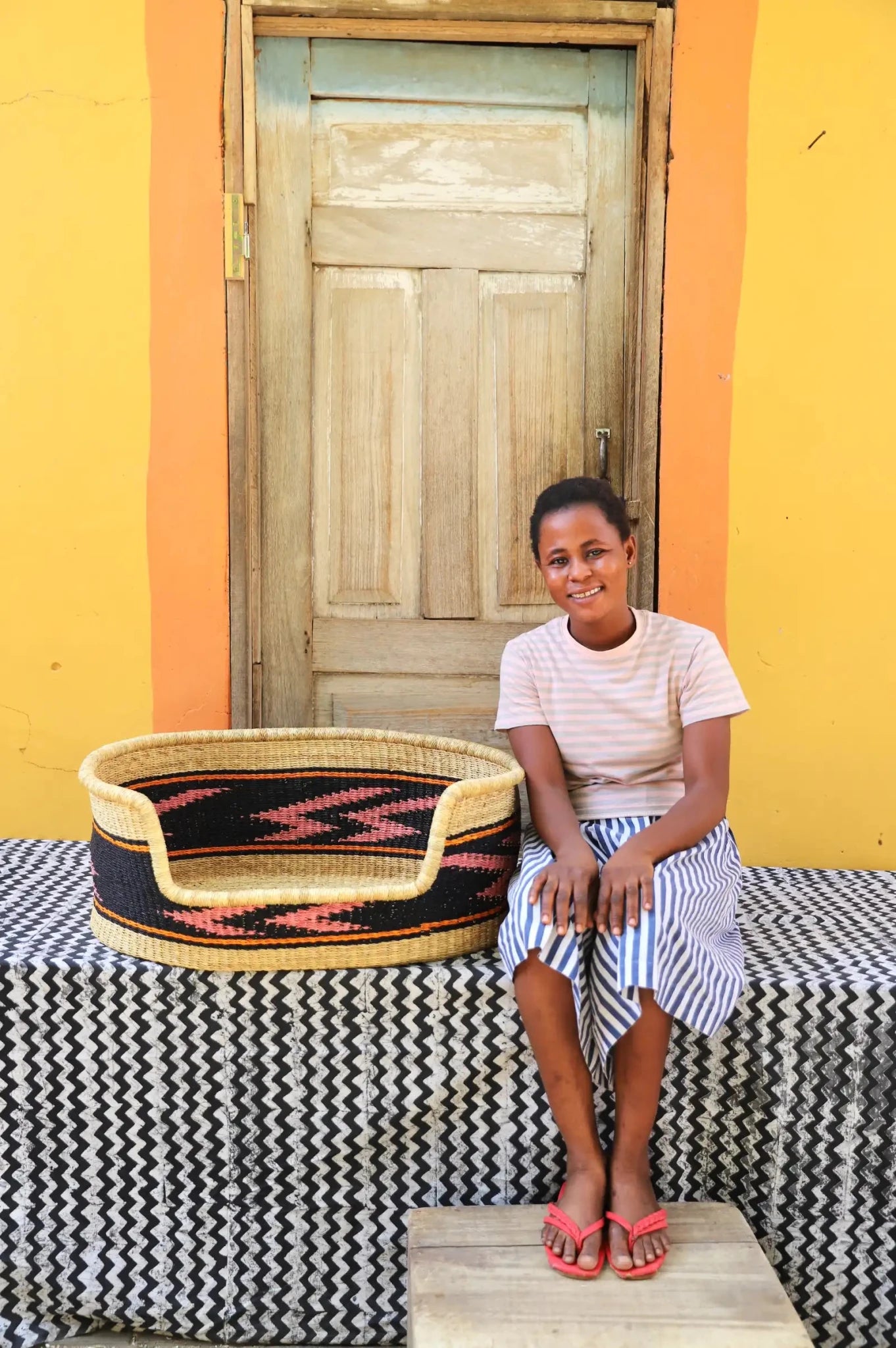 Woman  sitting on a bench with a woven basket next to a wooden door and yellow wall.
