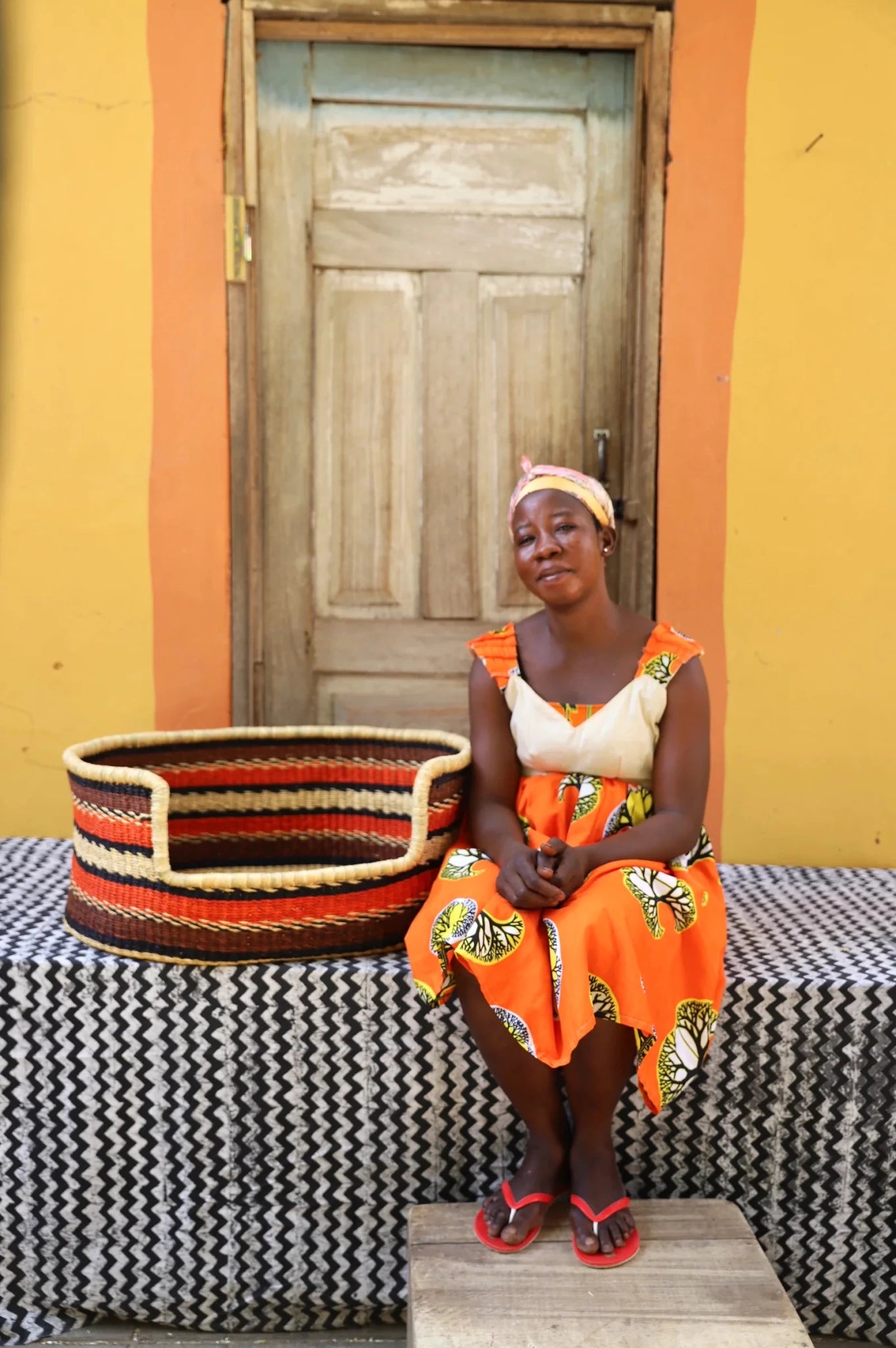 Woman sitting on a bench with a woven basket next to a wooden door and yellow wall.
