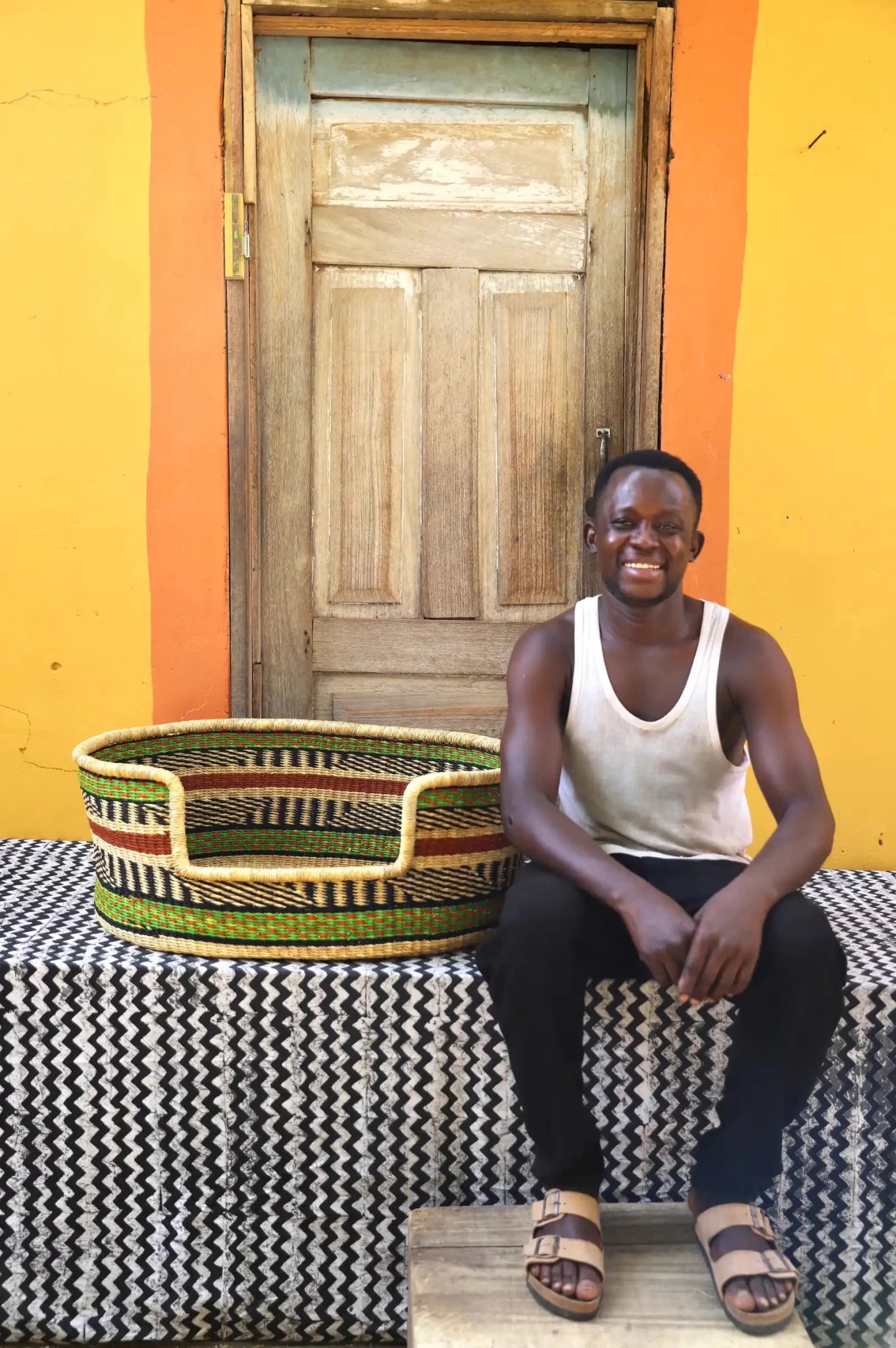 Man  sitting on a bench with a woven basket next to a wooden door and yellow wall.