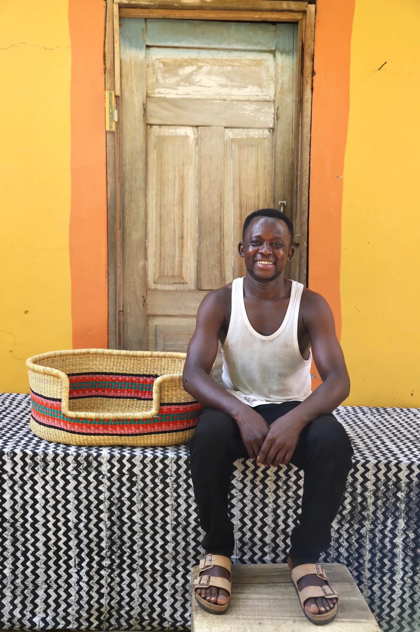 Woman/ Man  sitting on a bench with a woven basket next to a wooden door and yellow wall.
