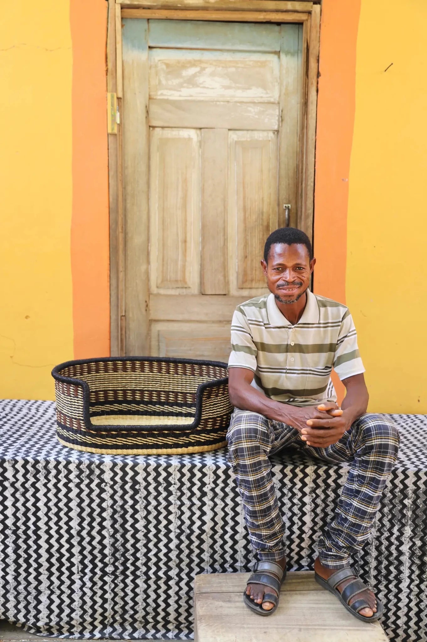 Man  sitting on a bench with a woven basket next to a wooden door and yellow wall.