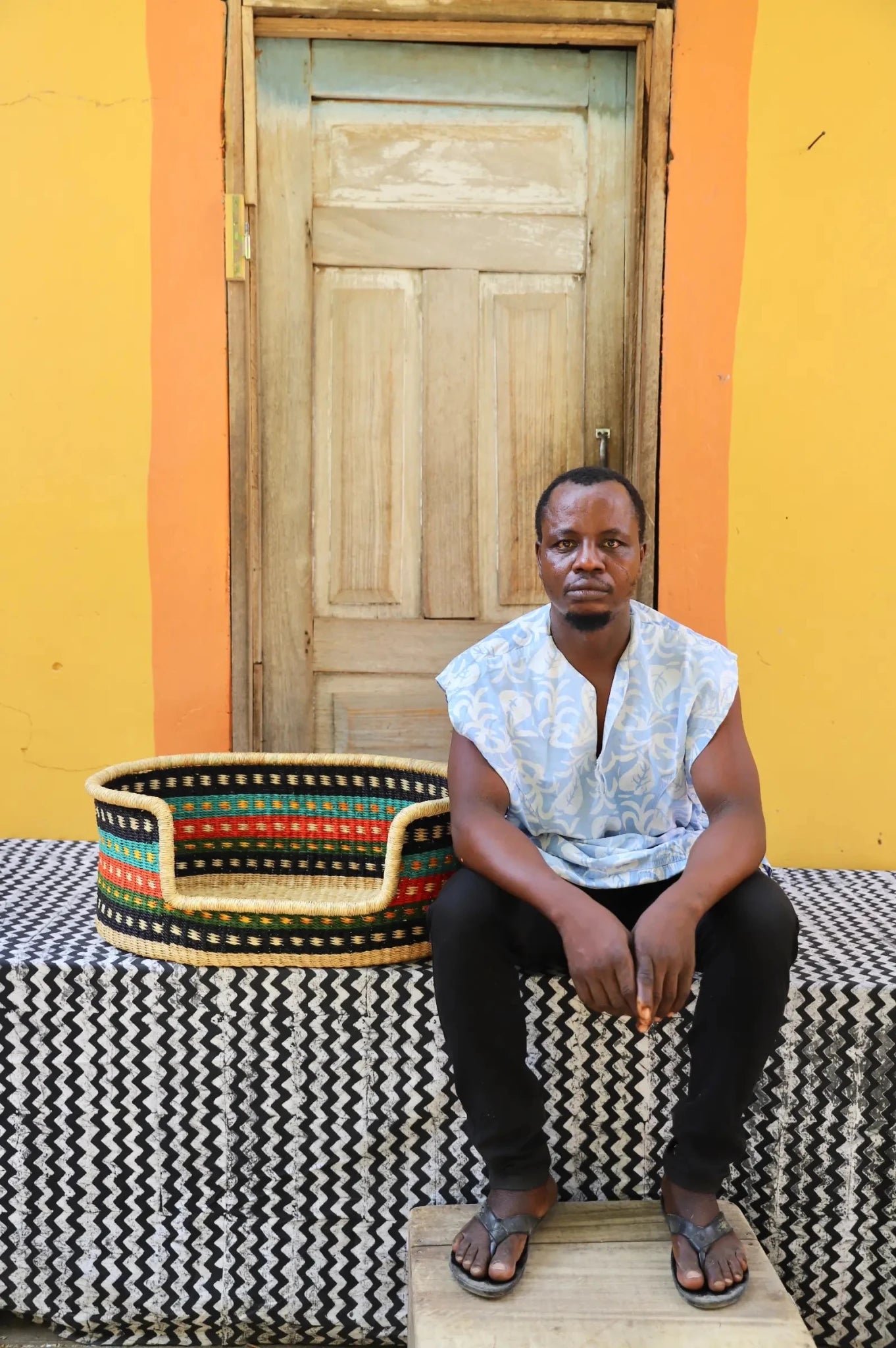 Man  sitting on a bench with a woven basket next to a wooden door and yellow wall.
