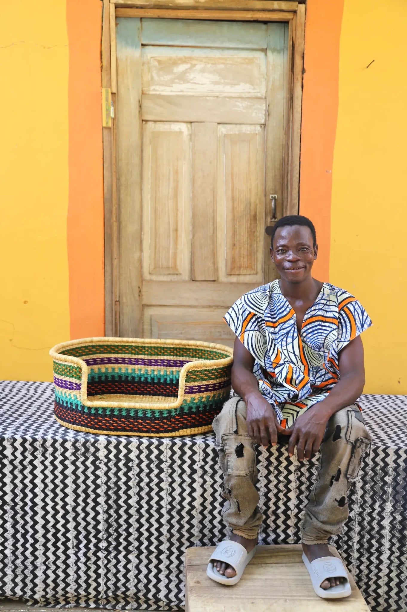 Woman/ Man  sitting on a bench with a woven basket next to a wooden door and yellow wall.