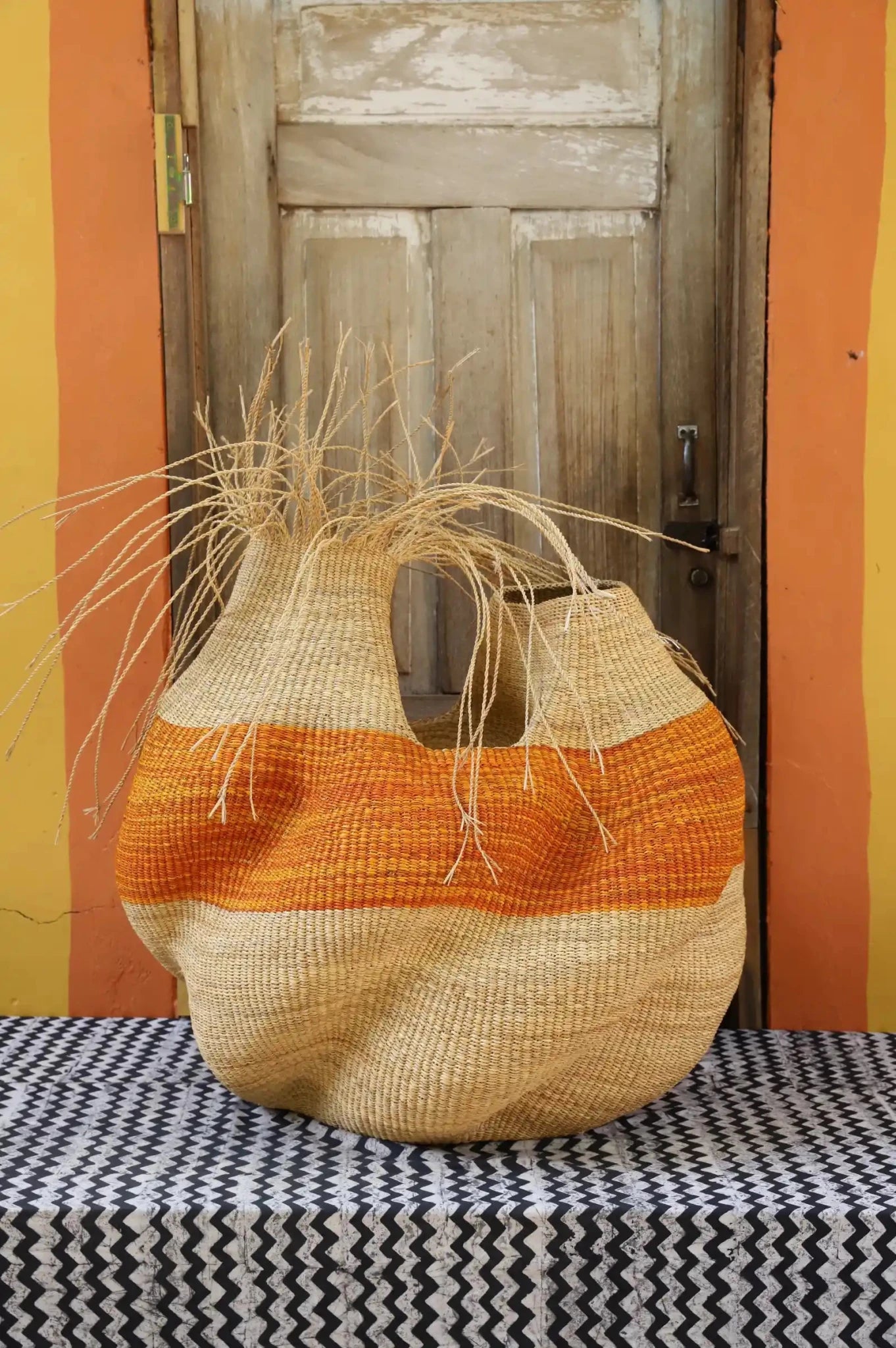 A colorful, handwoven piece sitting on a black and white cloth with decorative straw branching out against a wooden door