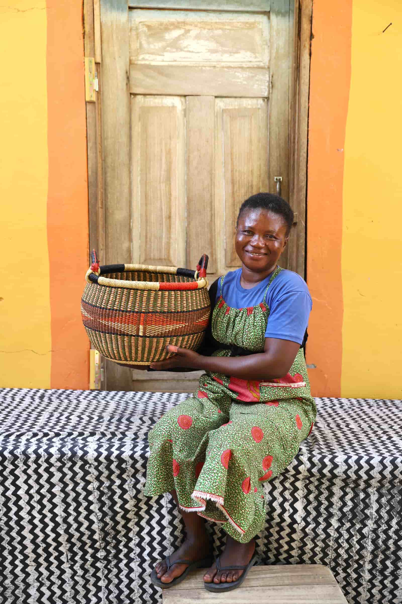 Artisan holding a colorful handwoven storage basket sitting on a black and white cloth against a wooden door.