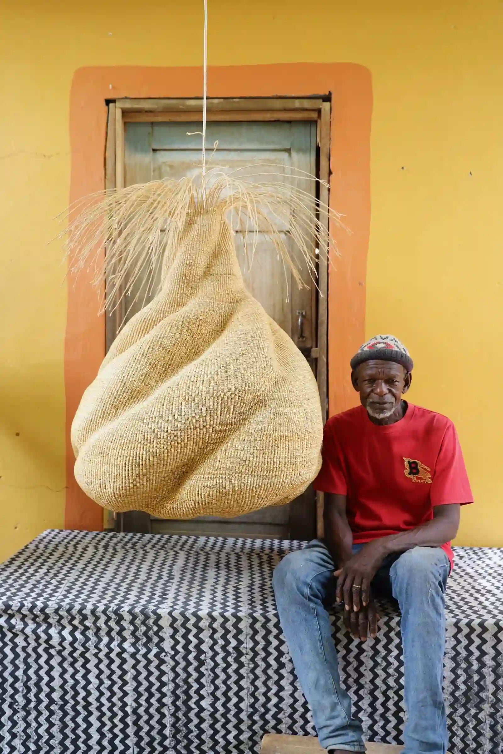 Man seated beside a Helix Pendant Lampshade in natural straw with a twisted form and straw fringe, before a wooden door.