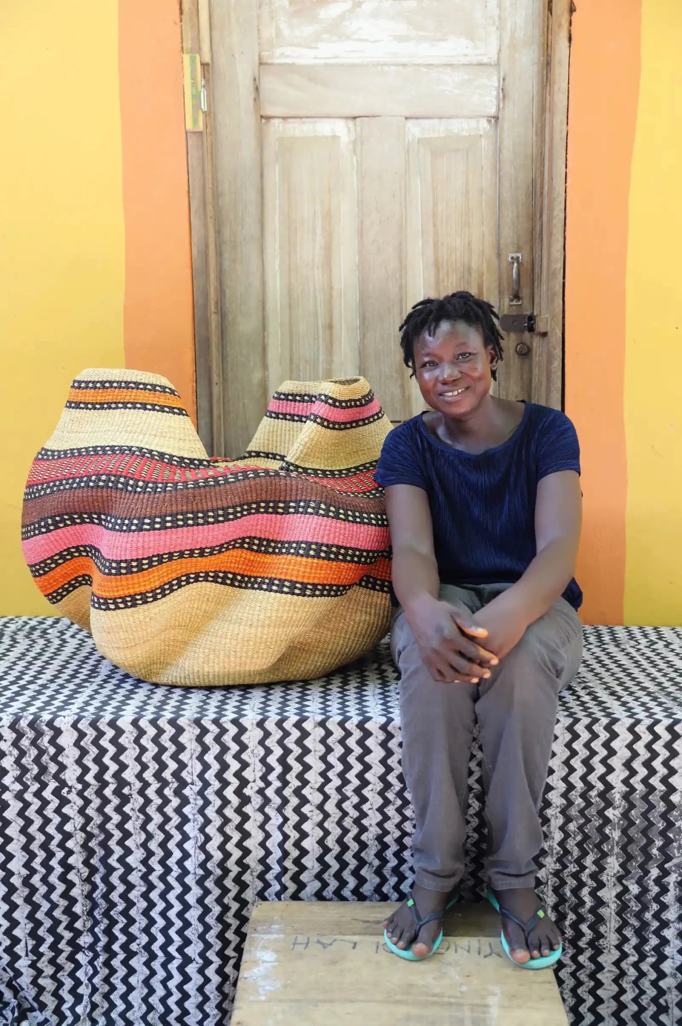 Woman sitting on a bench with a colorful woven basket next to her against a yellow wall.