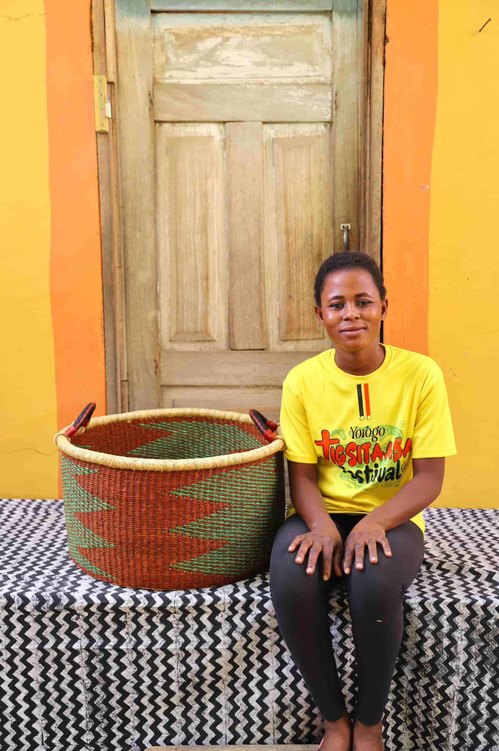 Woven Tub (Large) with woman weaver handwoven in Ghana sitting on black and white cloth and wooden door in background