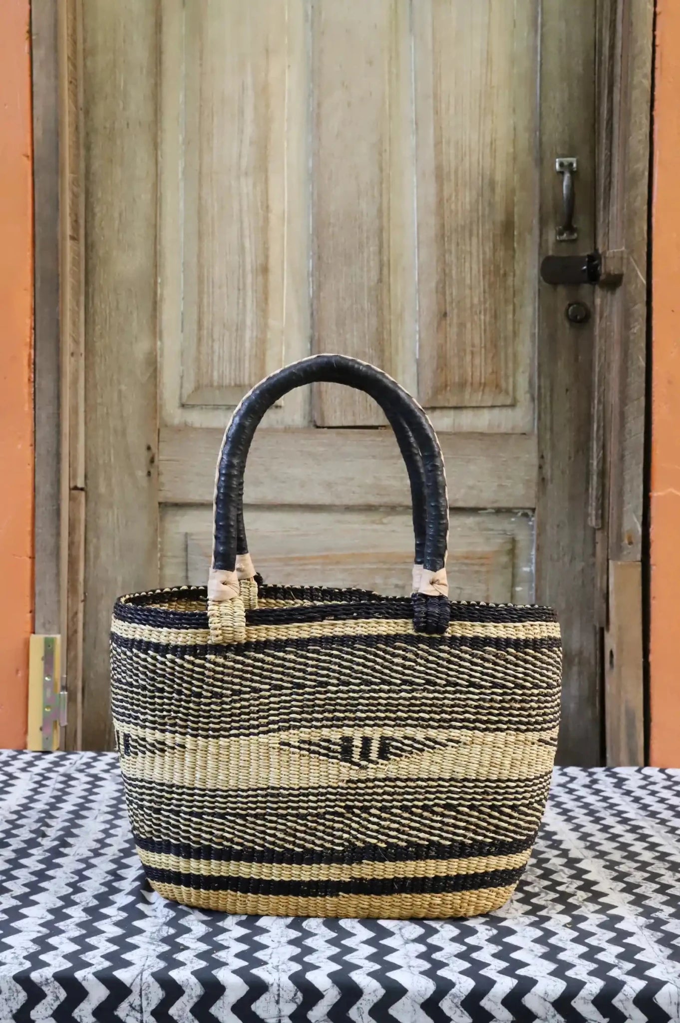 Tapoh carry basket in natural straw with dark navy bands and arrow motifs, dark leather handles, against a wooden door.