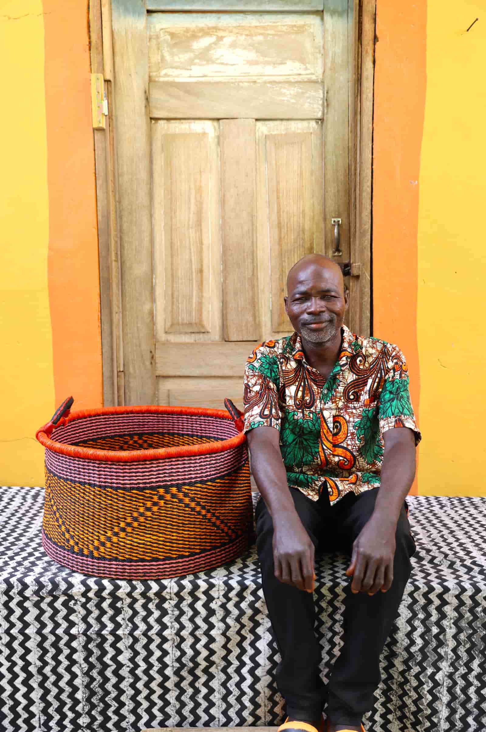 Woven Tub (Large) with man weaver handwoven in Ghana sitting on black and white cloth and wooden door in background

