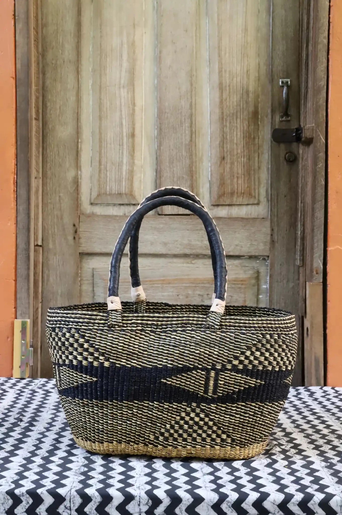 Tapoh carry basket in navy and natural straw with diamond and cross motifs, dark leather handles, against a wooden door.