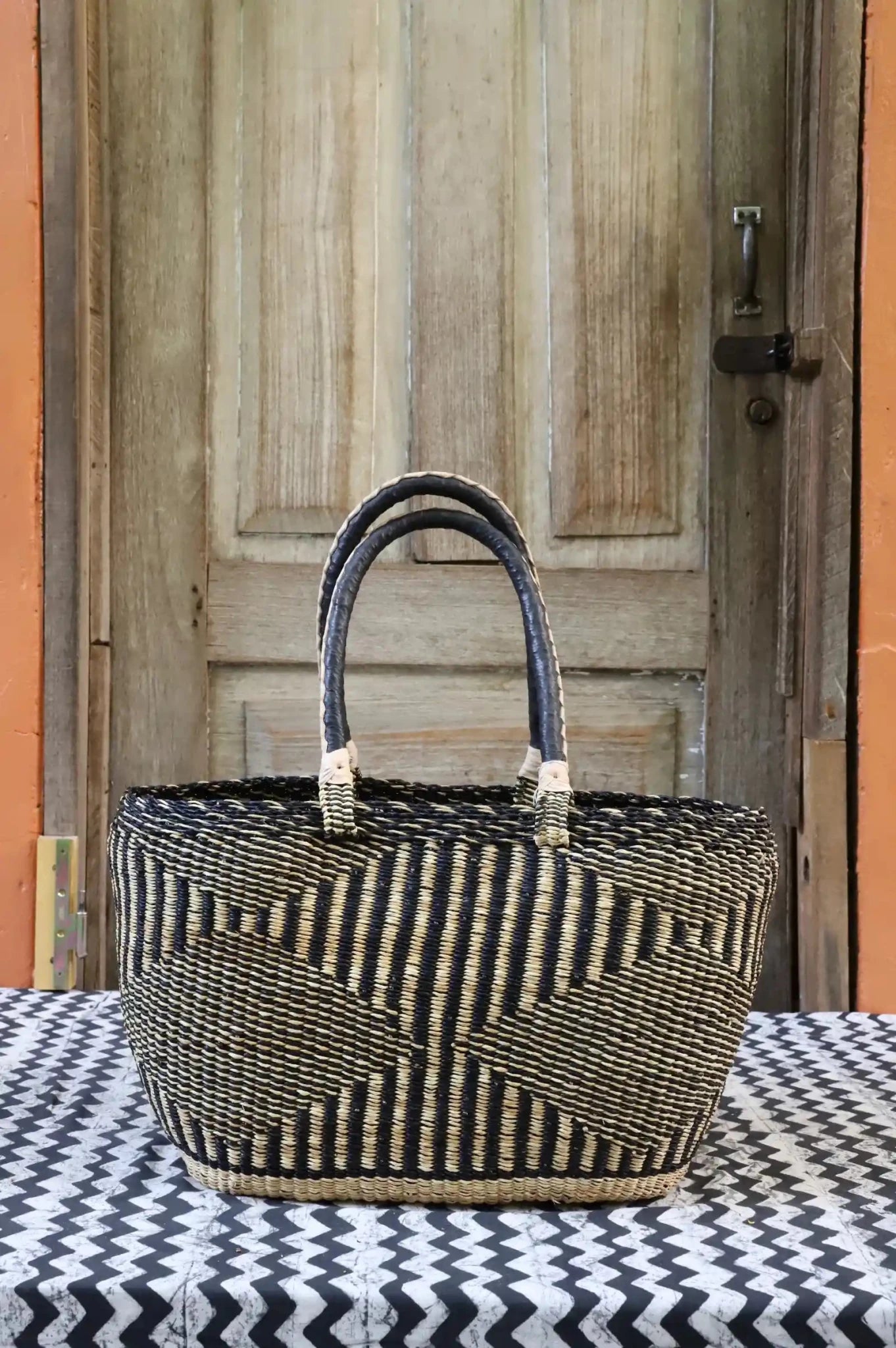 Tapoh carry basket in dark navy and natural straw vertical stripe weave, dark leather handles, against a wooden door.
