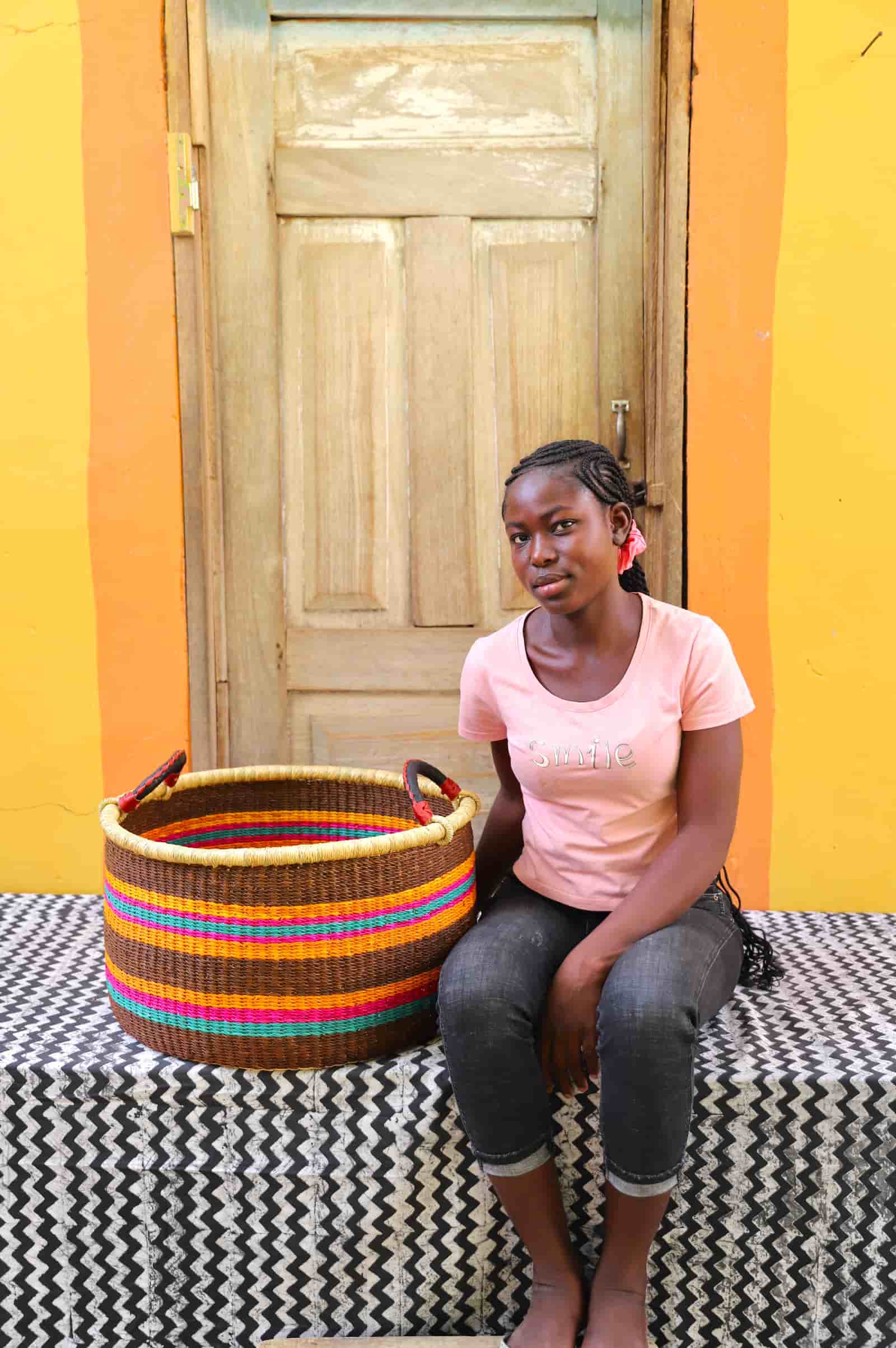Woven Tub (Medium) with woman weaver handwoven in Ghana sitting on black and white cloth and wooden door in background


