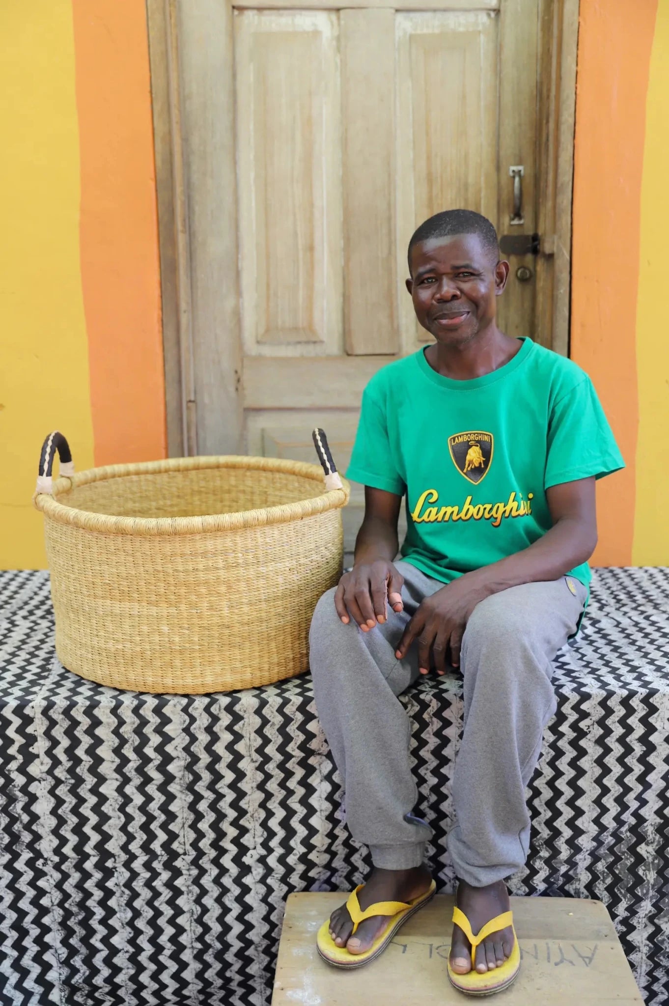 Woven Tub (Medium) with man weaver handwoven in Ghana sitting on black and white cloth and wooden door in background
