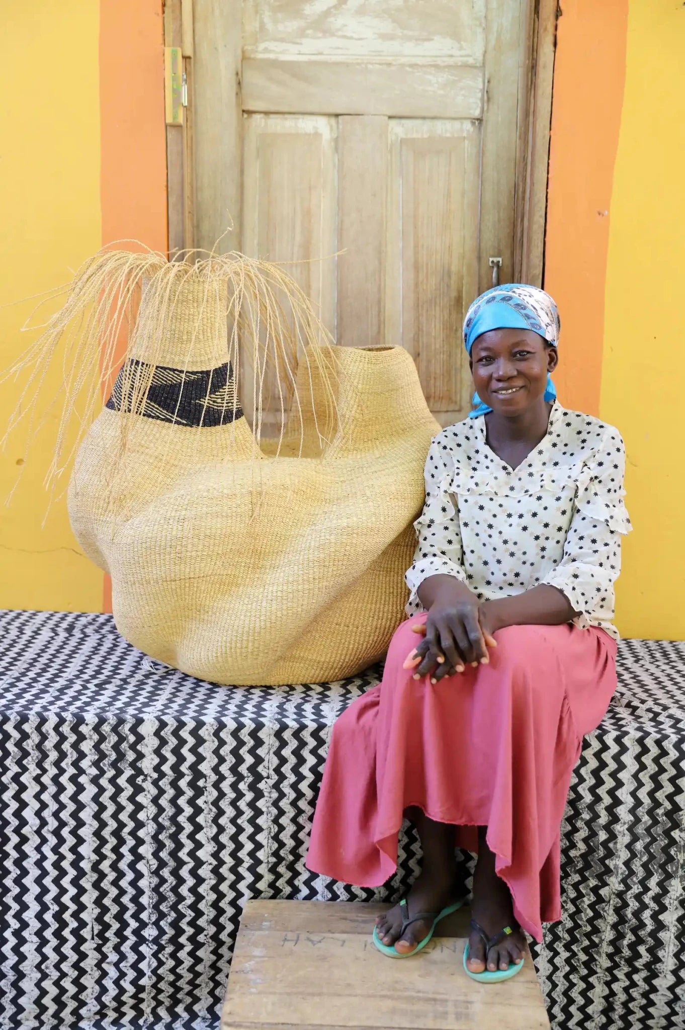 An artisan seated next to a handcrafted decorative basket with stylish straw branching out on a black and white cloth against a wooden door.
