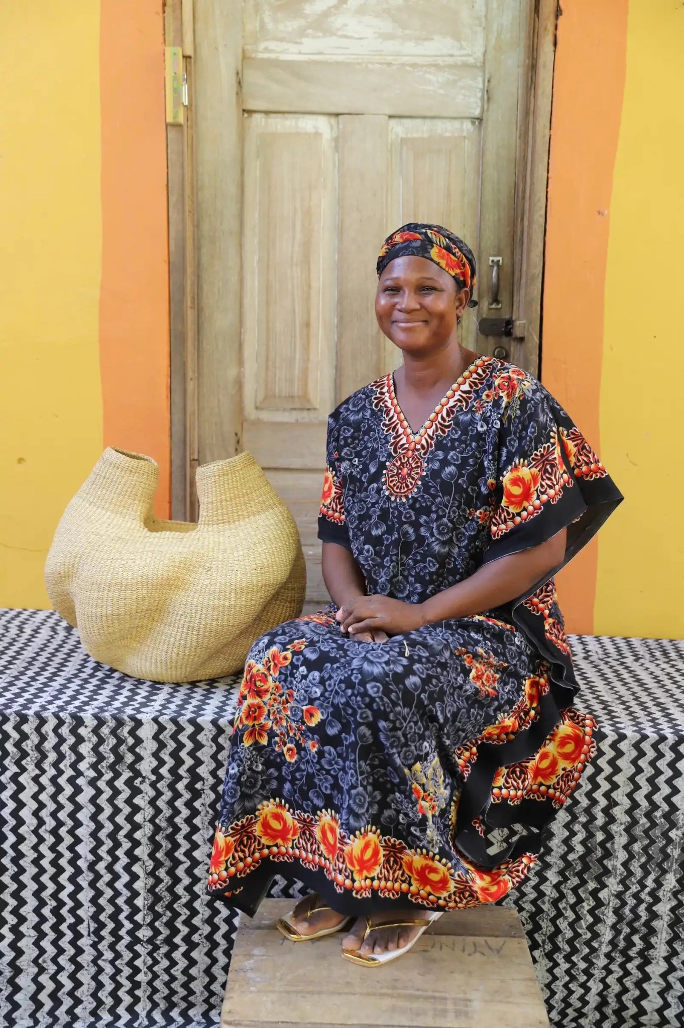 An artisan seated next to a handcrafted decorative basket on a black and white cloth against a wooden door.