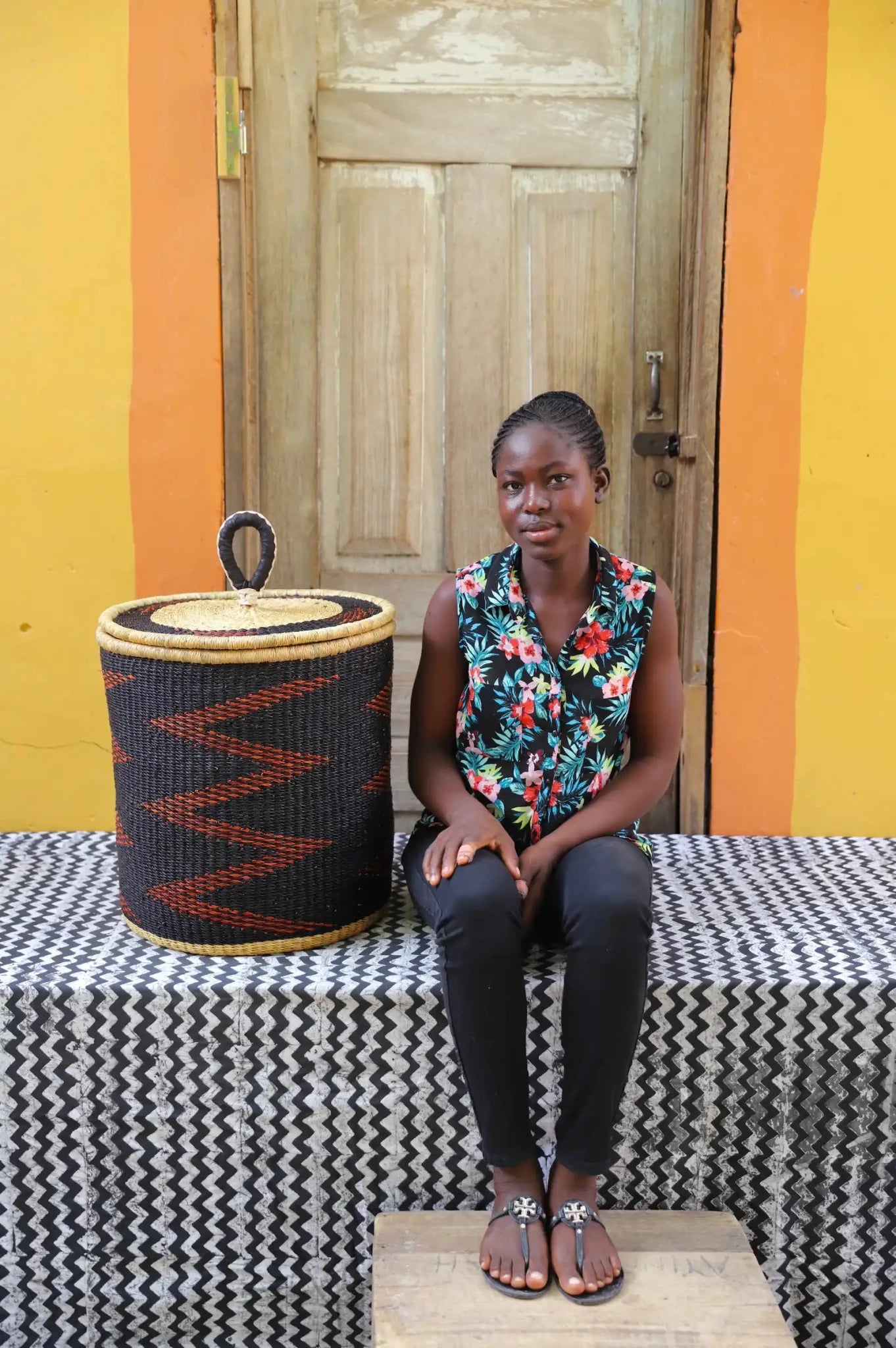 Woman sitting on a bench with a woven basket next to her against a colorful wall.