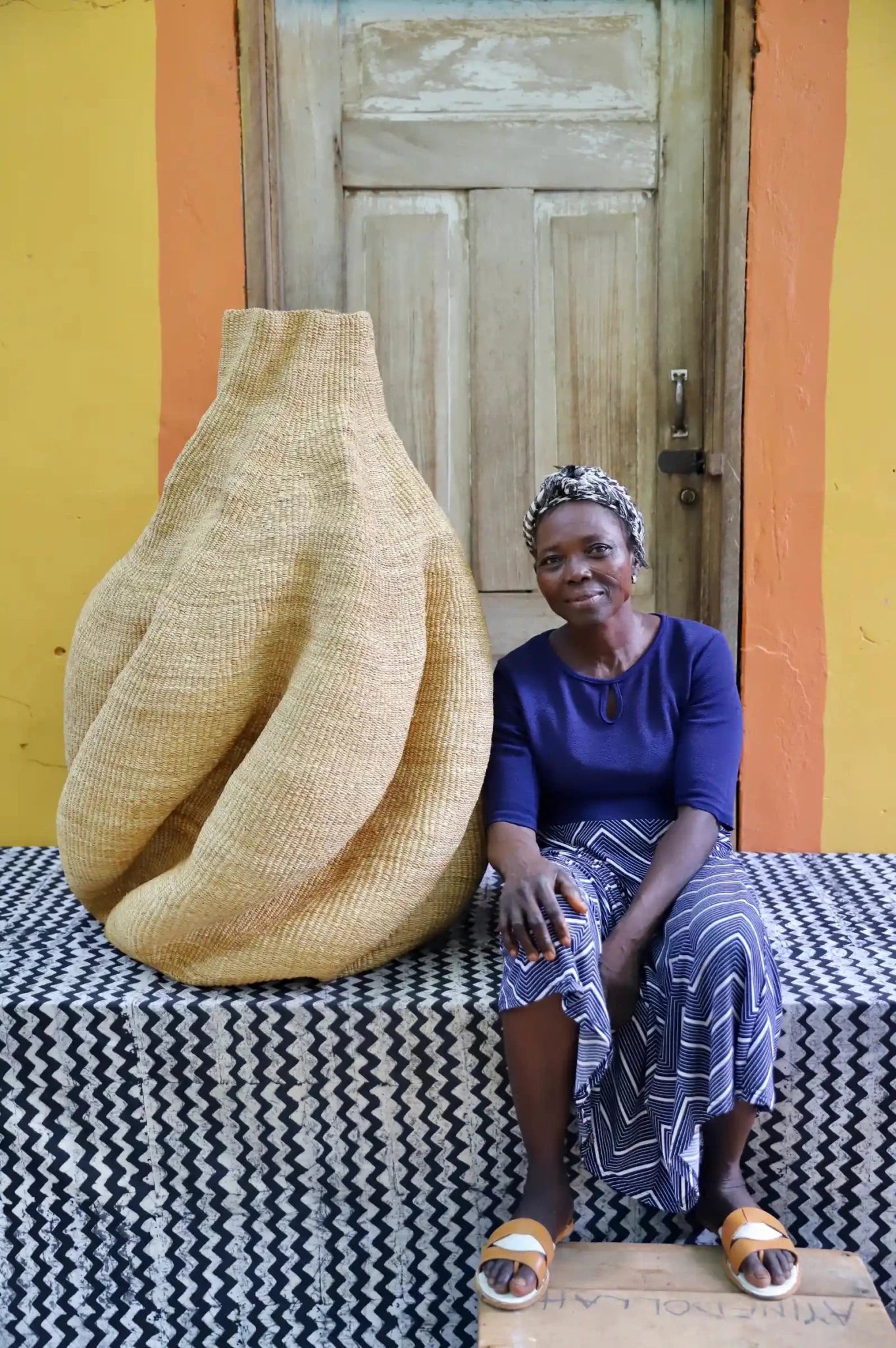 An artisan seated next to a handcrafted decorative basket  on a black and white cloth against a wooden door.