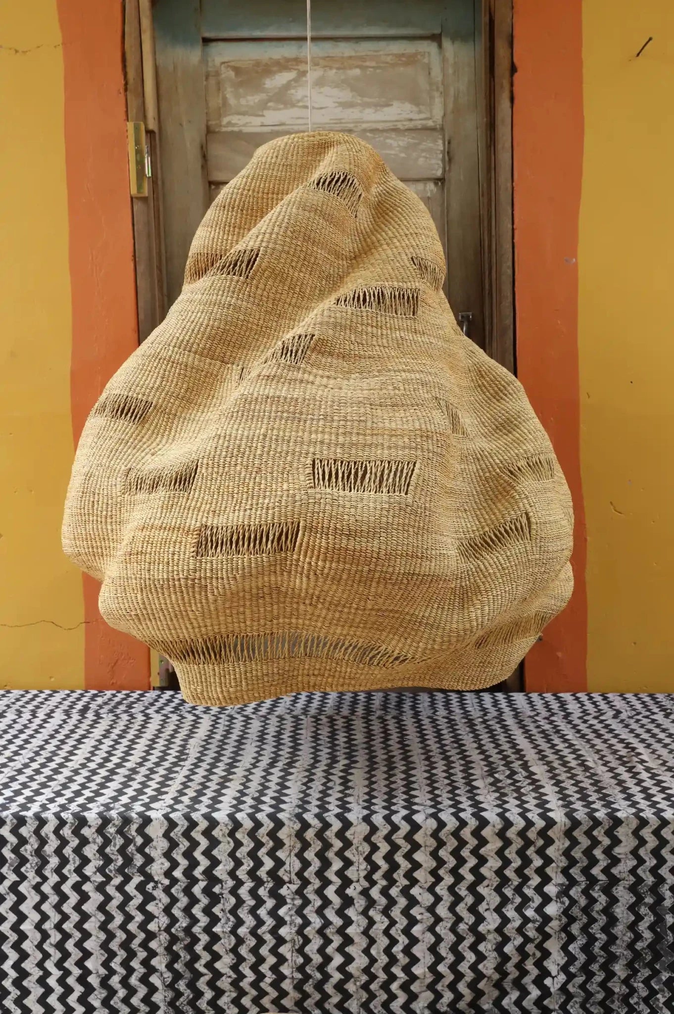 Textured beige bag on a black and white patterned surface with a yellow wall and wooden door in the background.