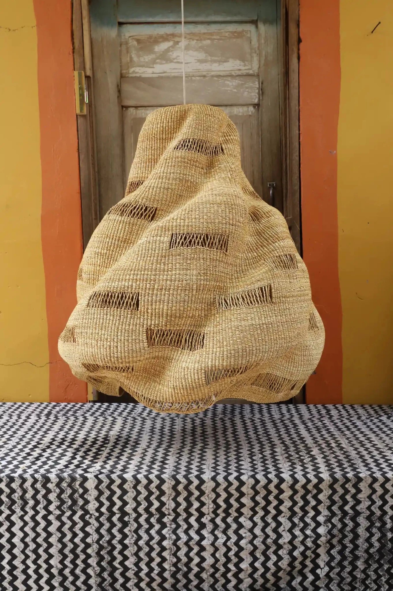 Woven bag hanging on a hook against a yellow wall with a black and white patterned surface.