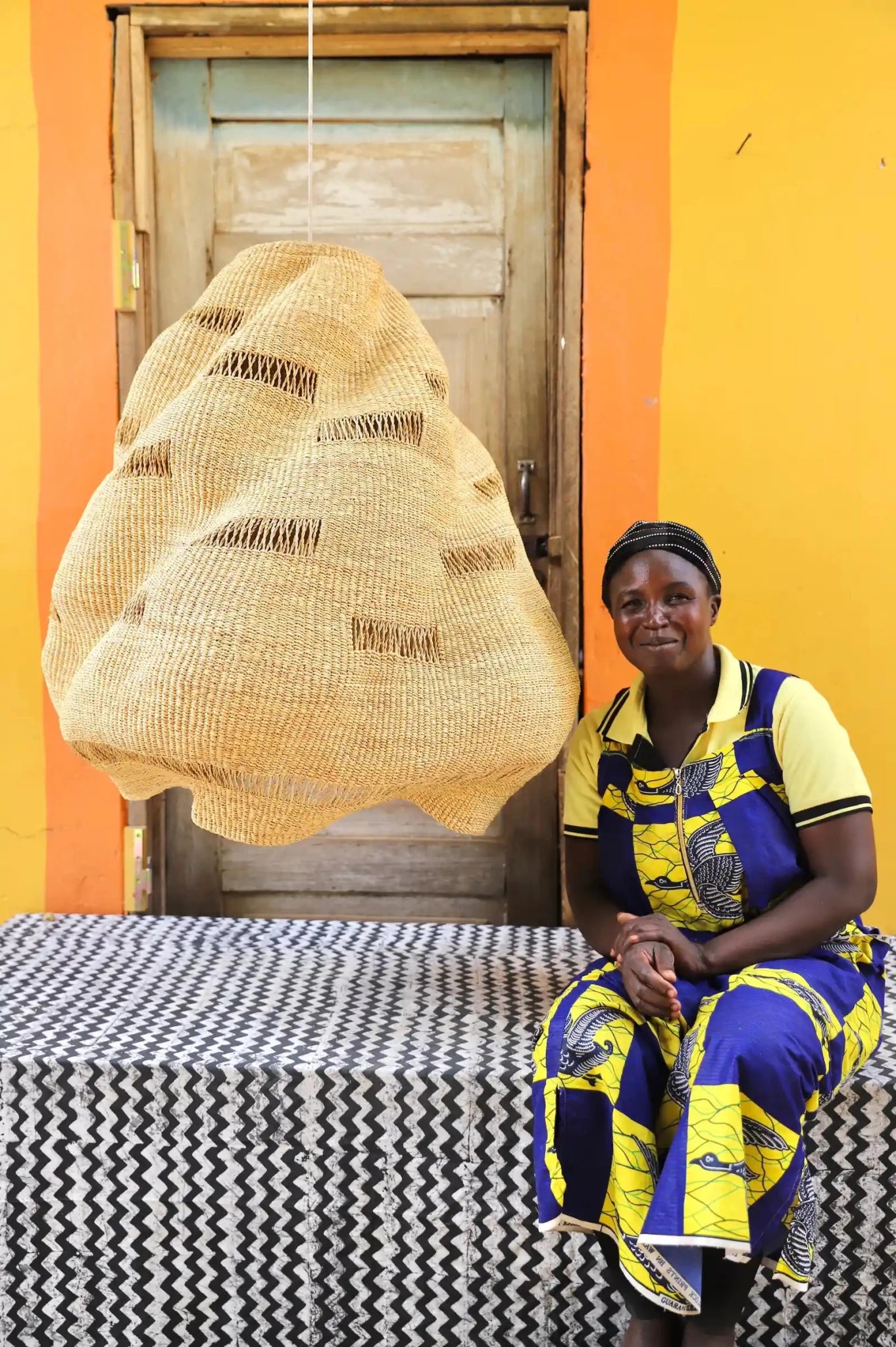 Woman in colorful traditional attire sitting on a bench with a woven basket in front of a wooden door and orange wall.