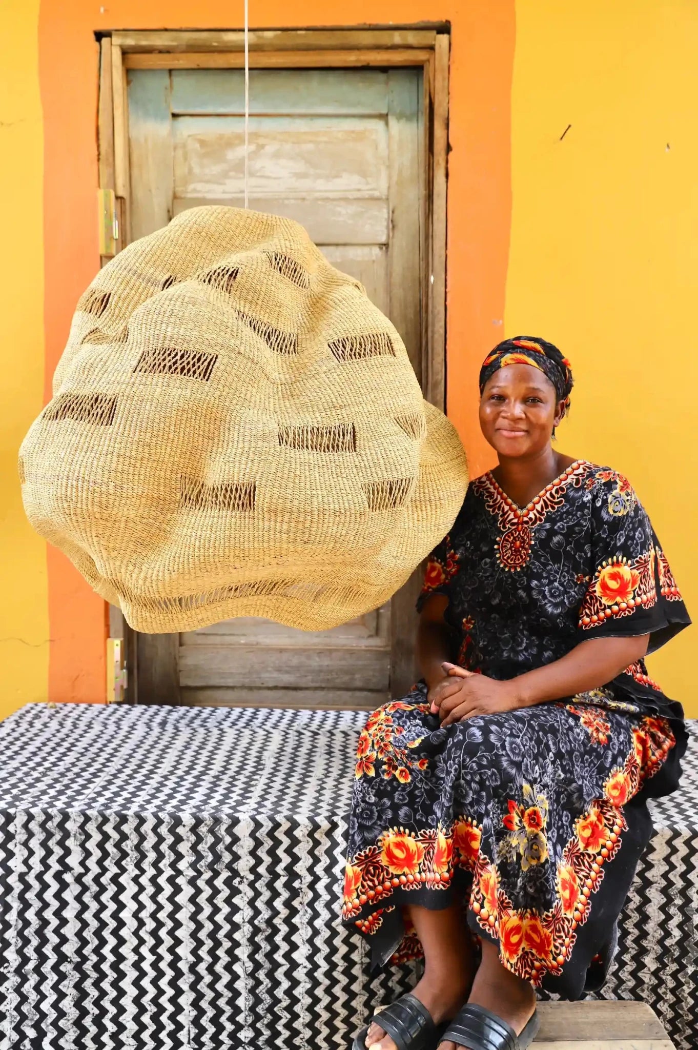 Woman in traditional attire sitting on a bench with a woven hat in front of a yellow wall.