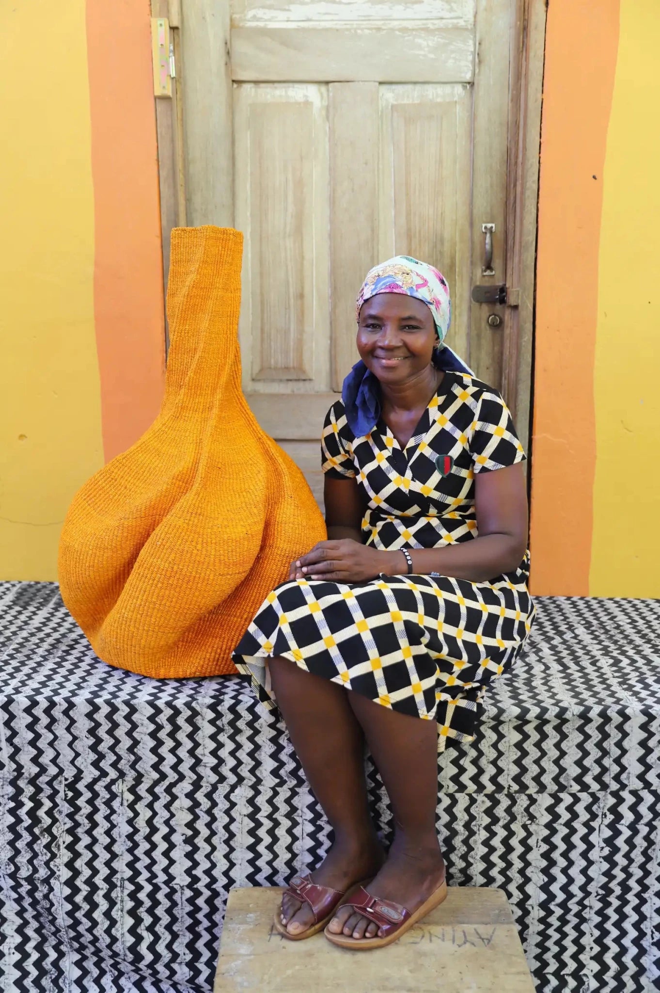 Woman sitting with yellow basket with decorative curves on black and white cloth in front of a wooden door