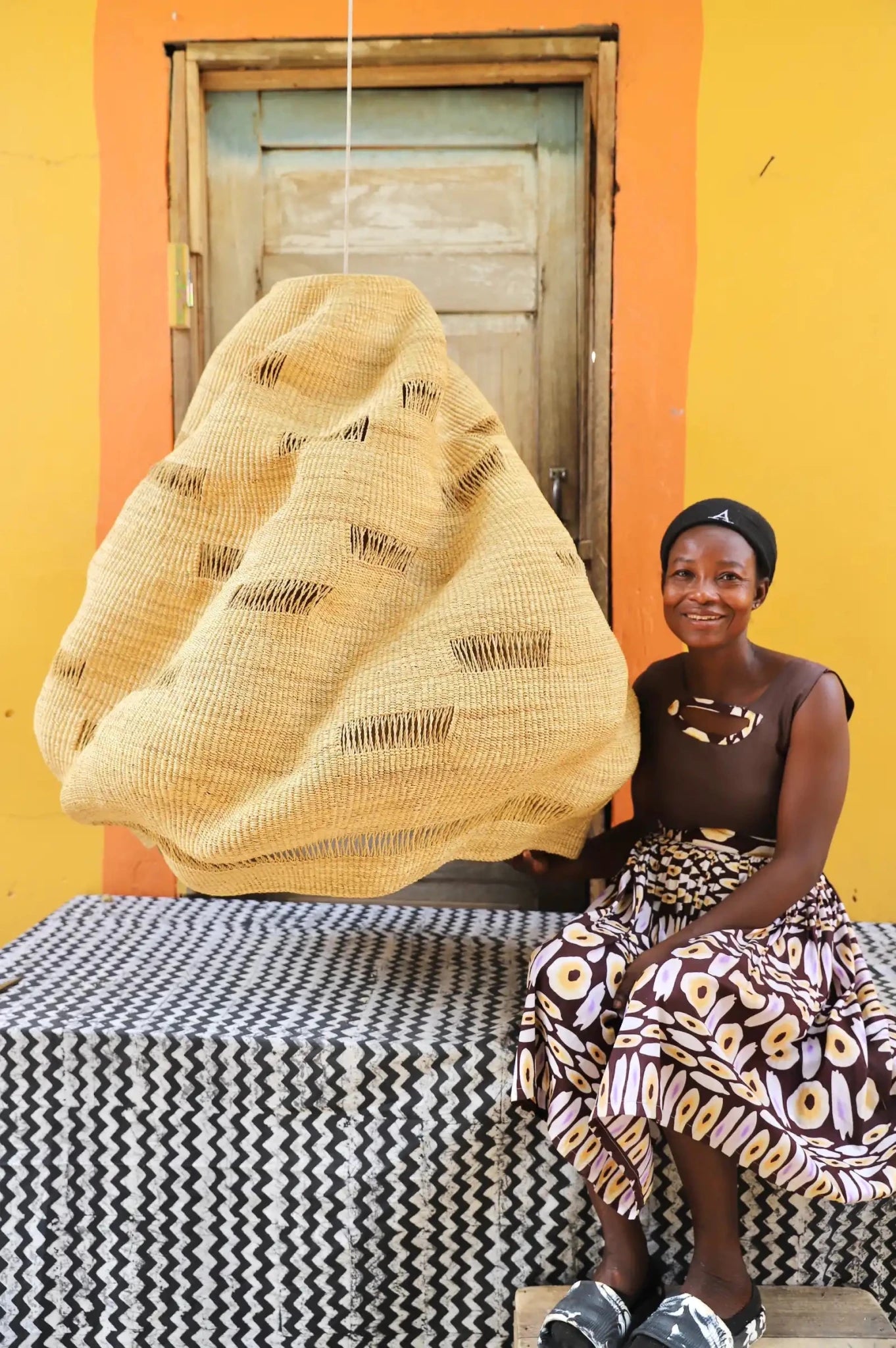 Woman sitting on a bench with a large woven basket against a yellow wall.