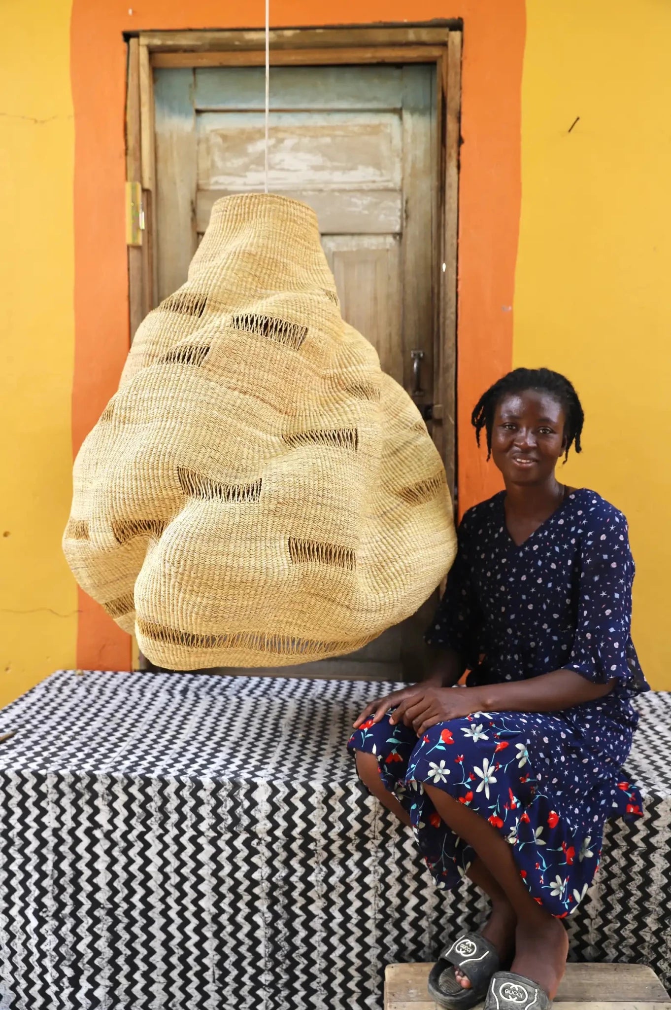 Woman sitting on a bench with a large woven basket behind her against an orange wall.