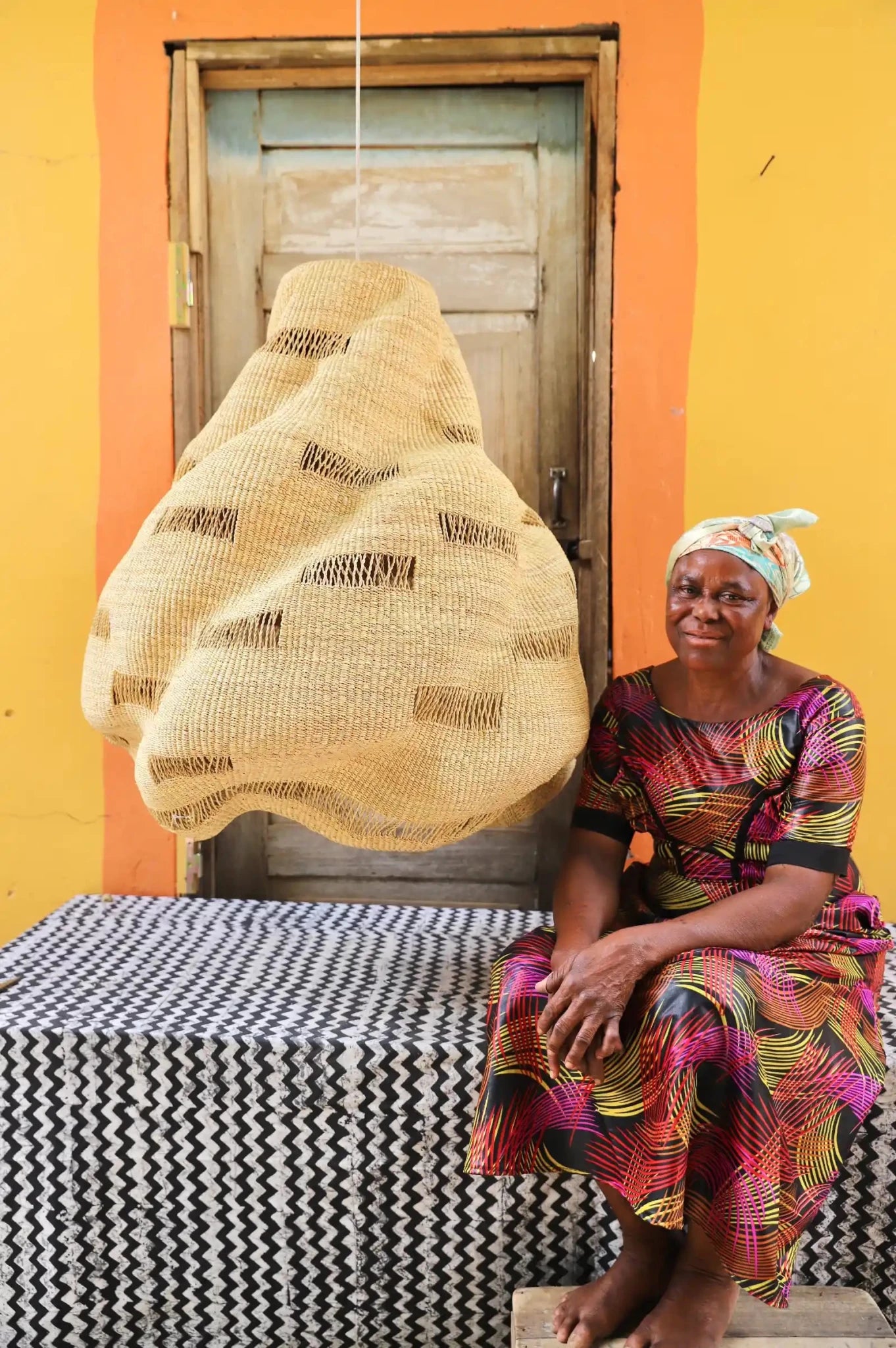 Woman in a colorful dress sitting on a bench with a woven basket against a yellow wall.