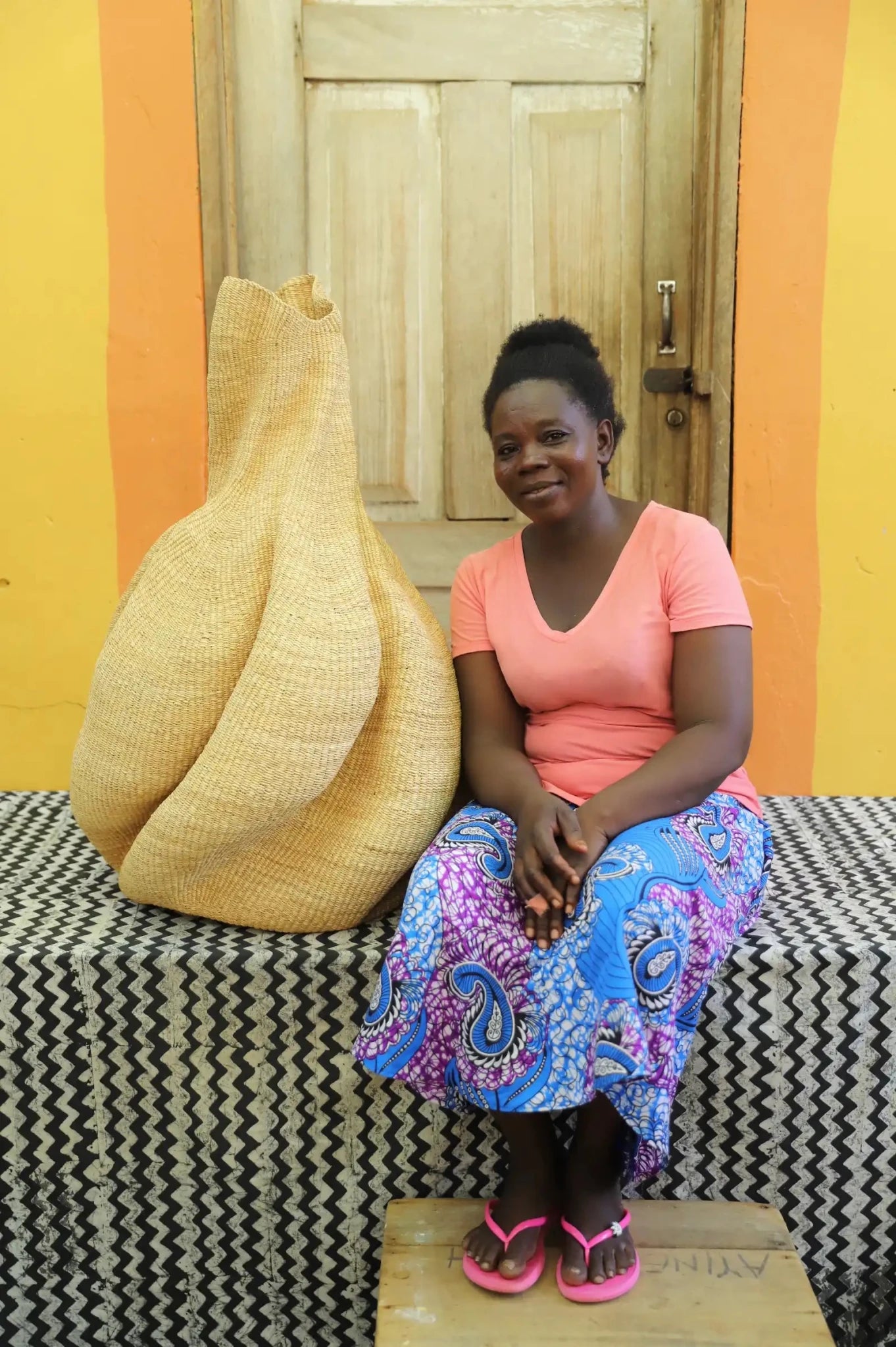 An artisan seated next to a handcrafted decorative basket  on a black and white cloth against a wooden door.