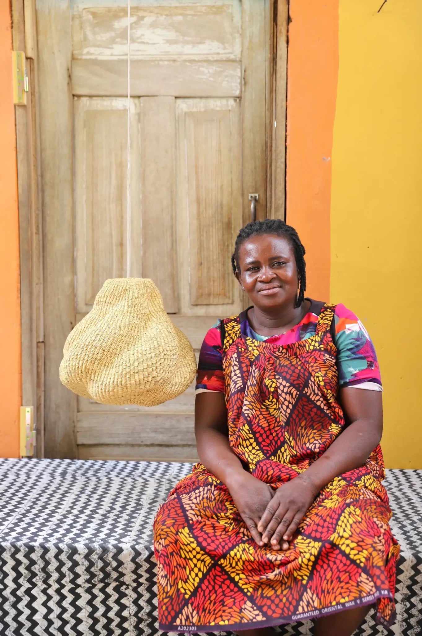 Woman in a colorful dress sitting on a bed with a woven basket in the background.
