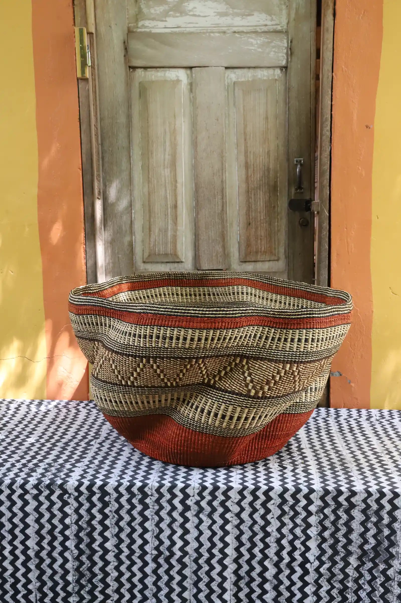 Large woven basket with geometric pattern on a black and white checkered tablecloth in front of a wooden door.