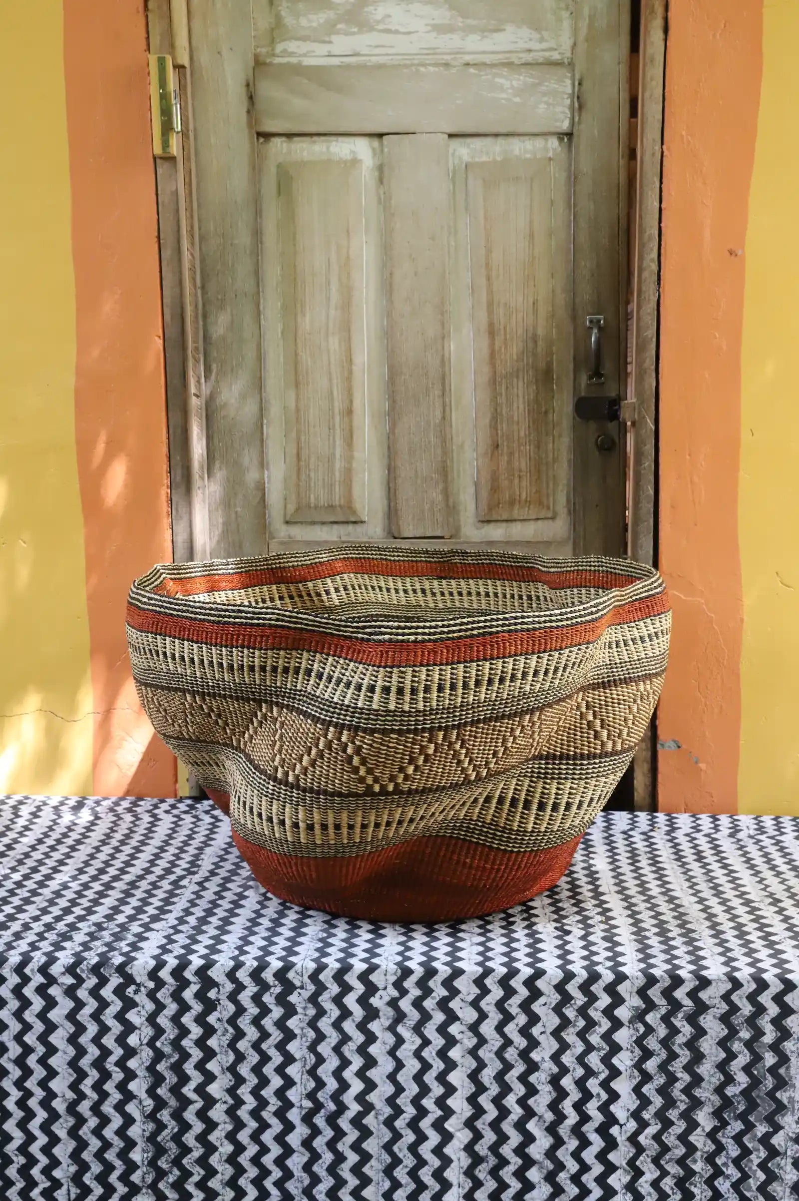 Large woven basket with geometric pattern on a black and white checkered tablecloth in front of a wooden door.