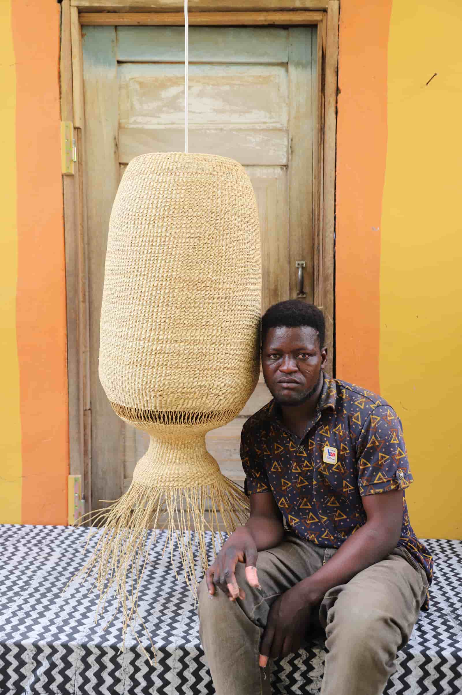 Man sitting on a black and white cloth  with a large woven basket above him against a colorful wall.