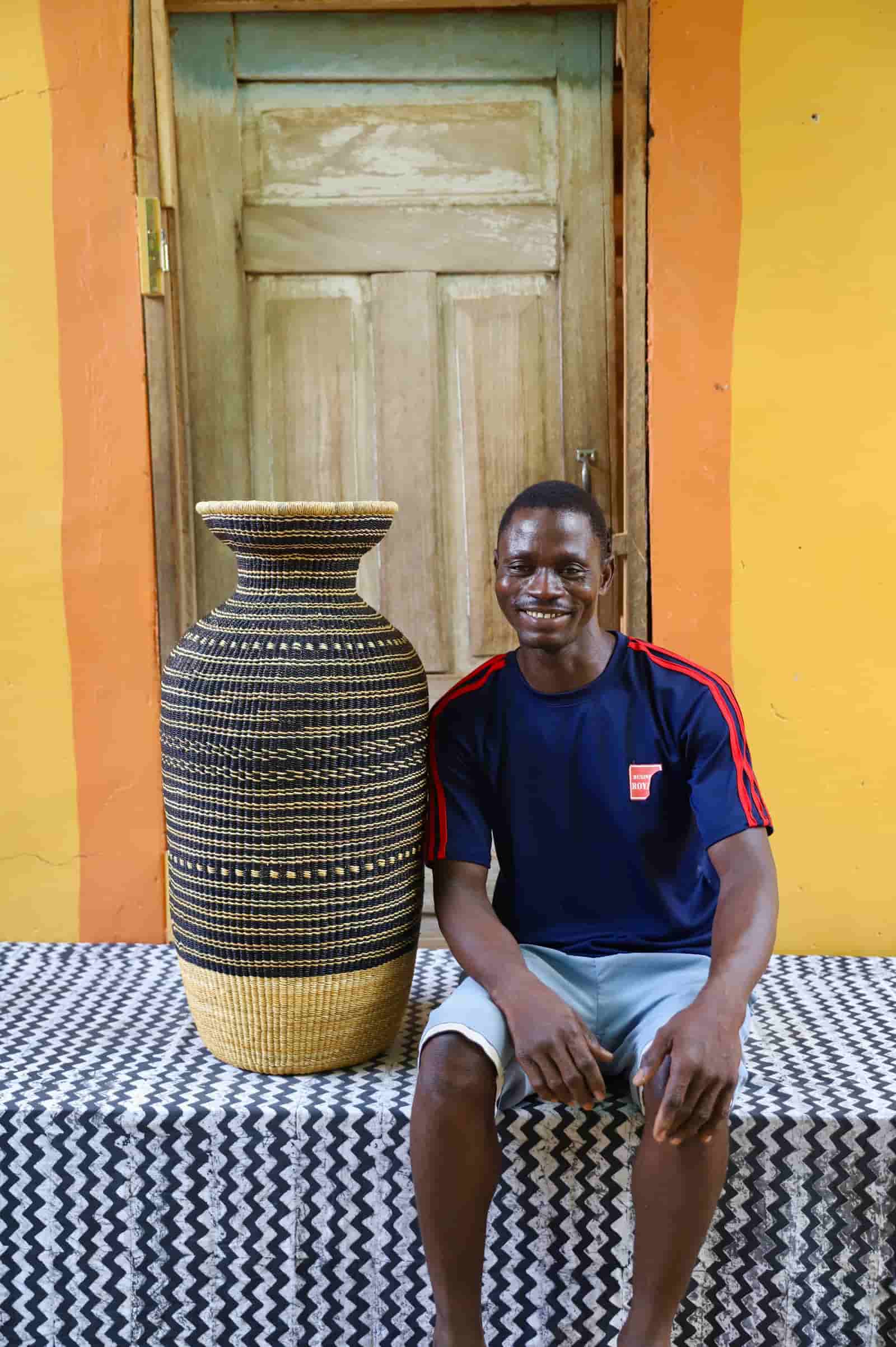 Man sitting next to a large woven décor basket on a black and white with a yellow wall and wooden door in the background