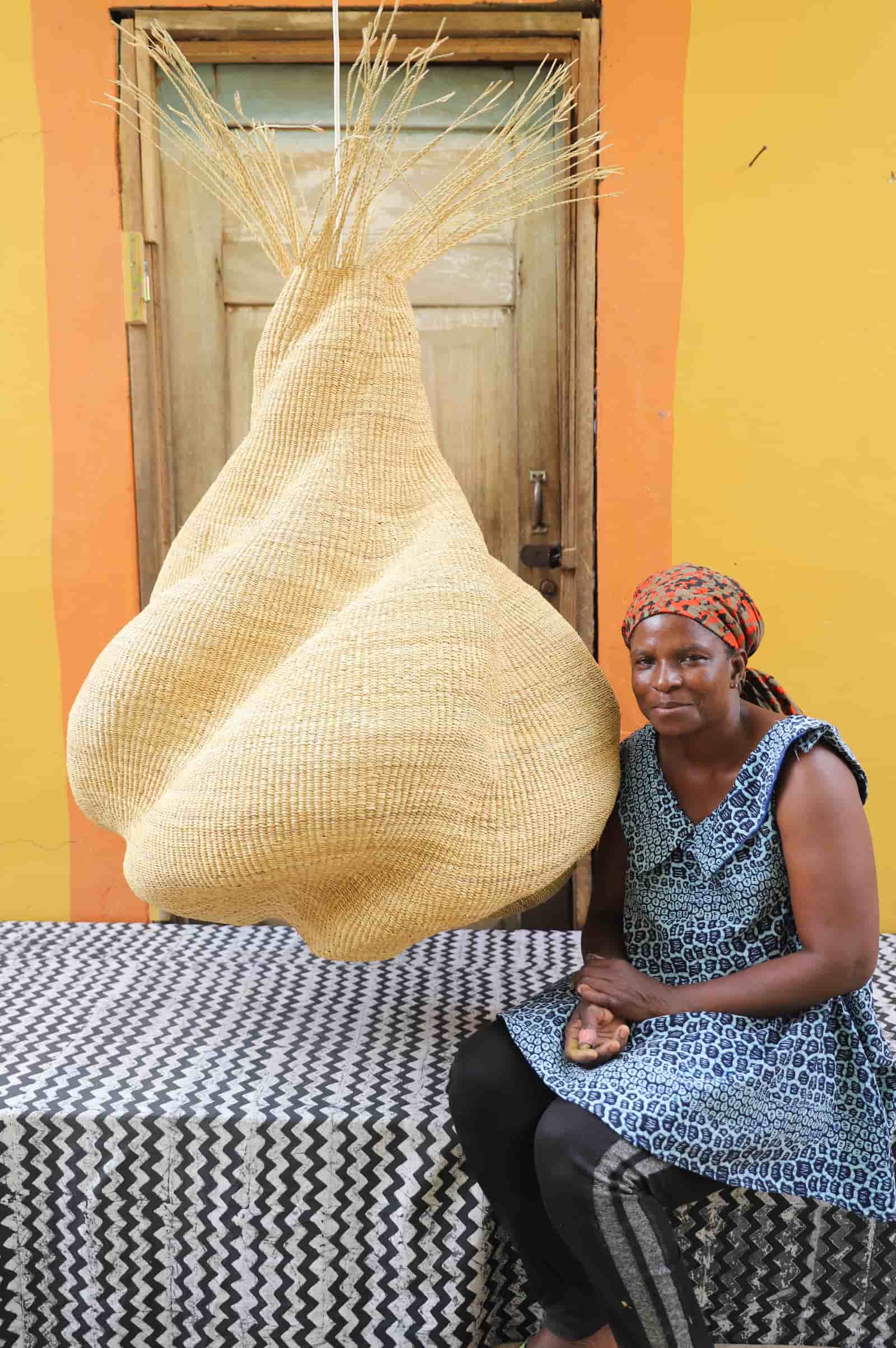 Woman sitting on a black and white cloth  with a large woven basket in front of a yellow wall.