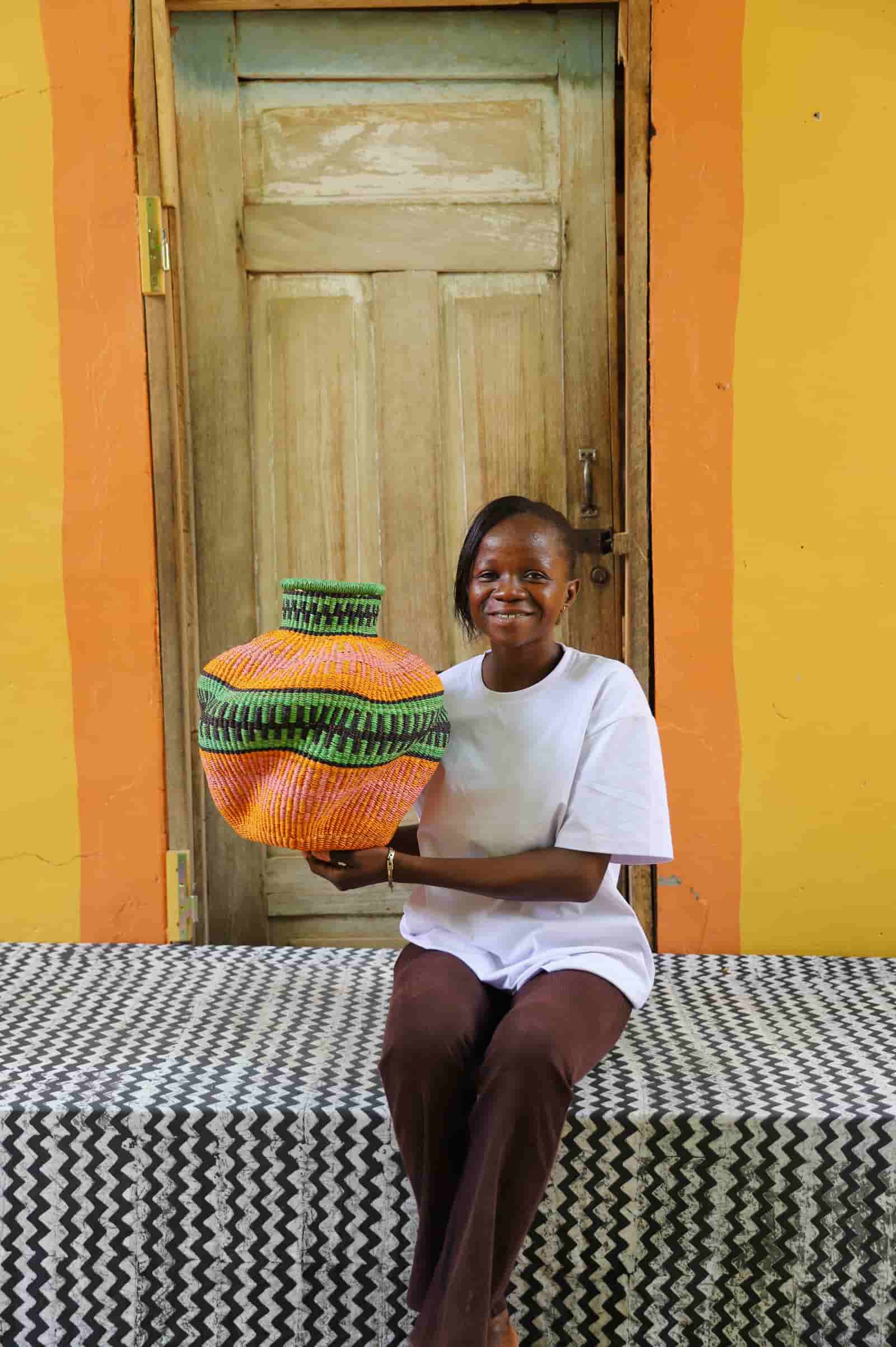 Woman sitting on a patterned bench next to a colorful woven basket against a yellow wall.