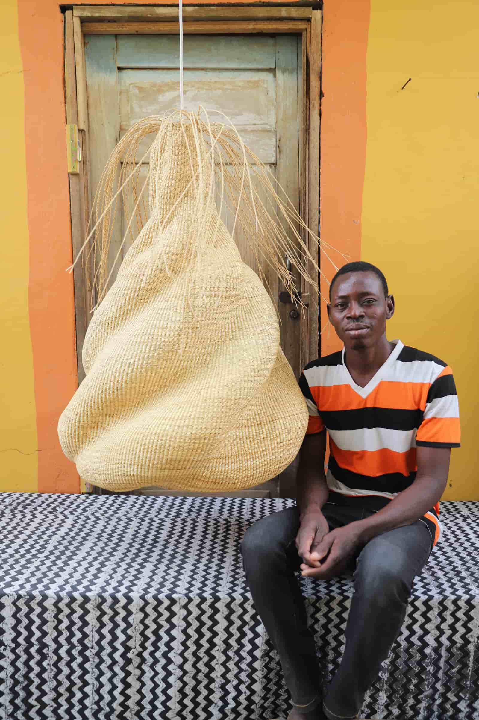 Man sitting on a black and white cloth  with a large woven basket behind him against a colorful wall.