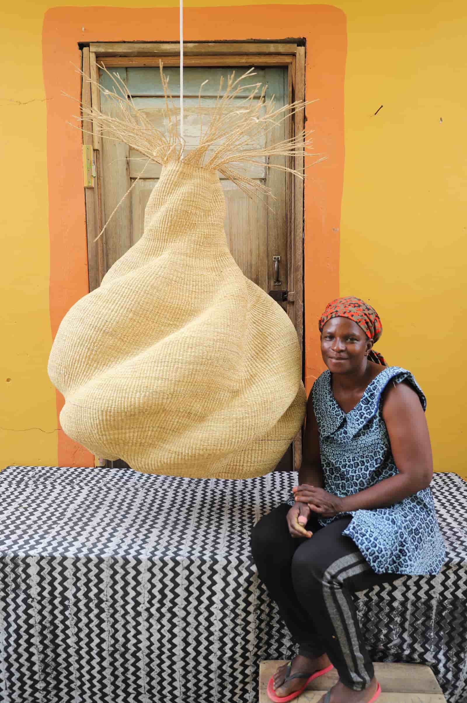 Woman sitting on a bench with a large woven basket in front of a yellow wall.