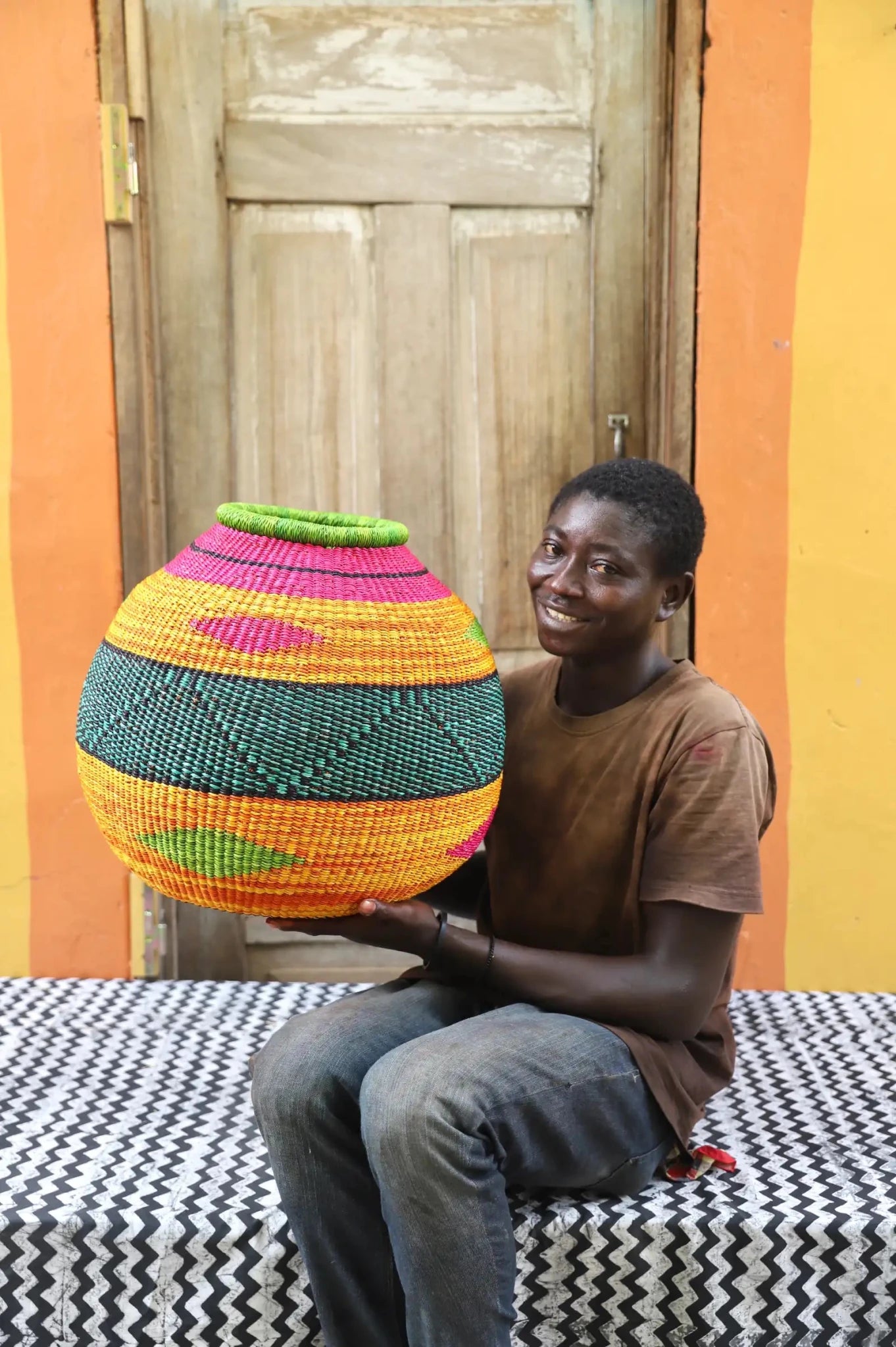 Person holding a colorful woven basket against a wooden door and orange wall.