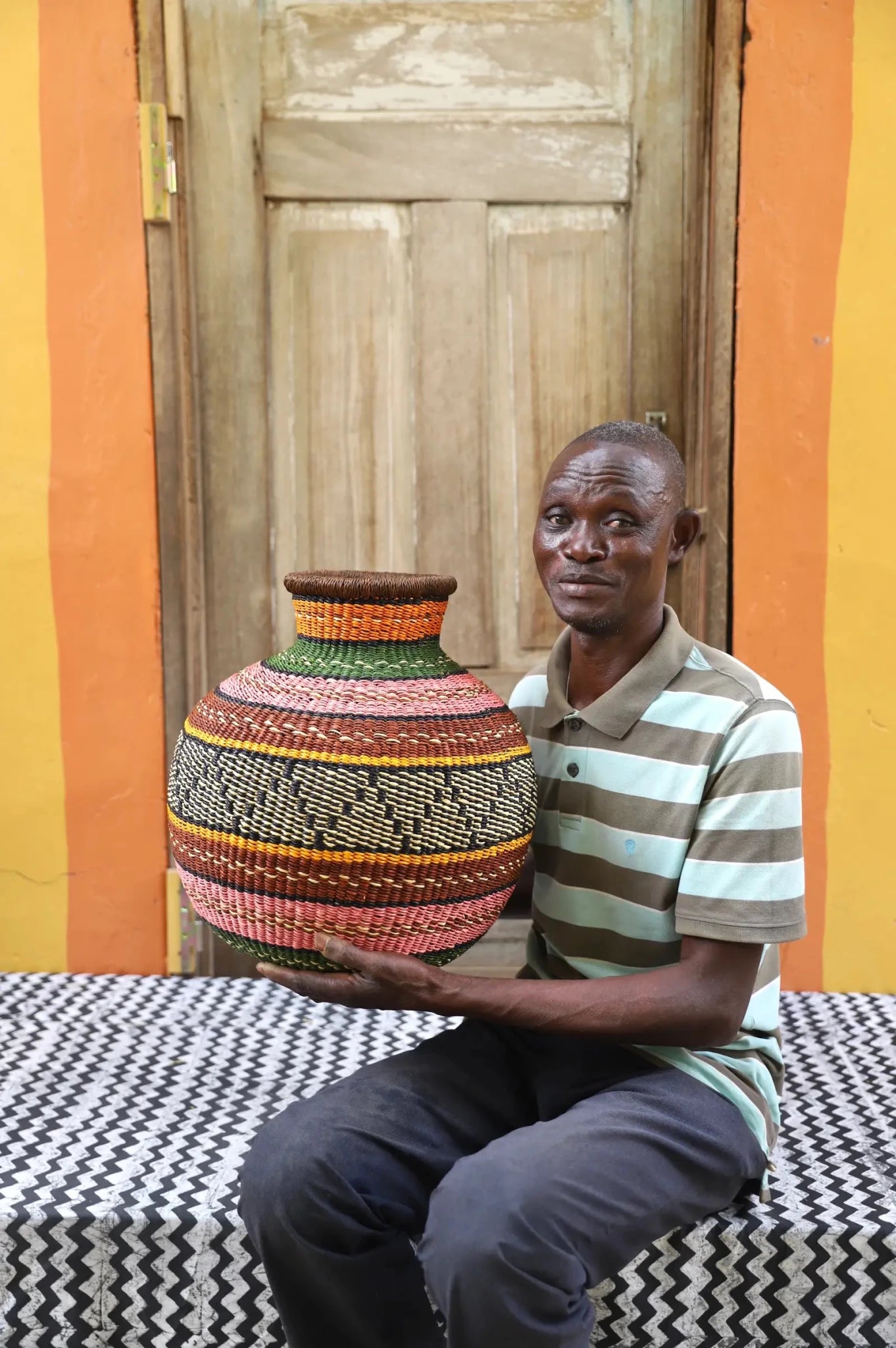 Artisan holding a colorful woven storage basket  on a black and white cloth against a wooden door.