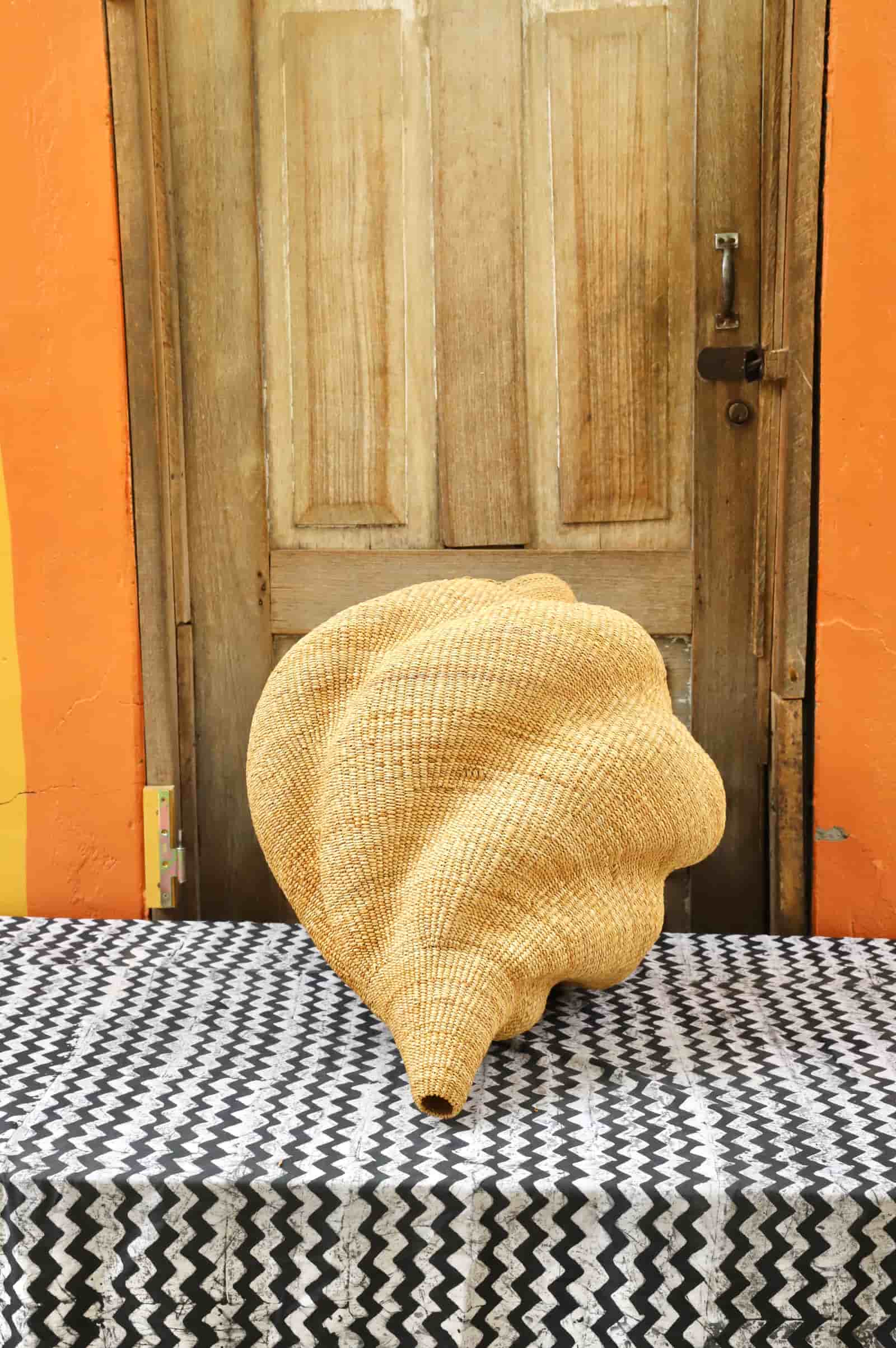 Woven conch  lampshade on a black and white patterned surface with an orange wall and wooden door in the background.