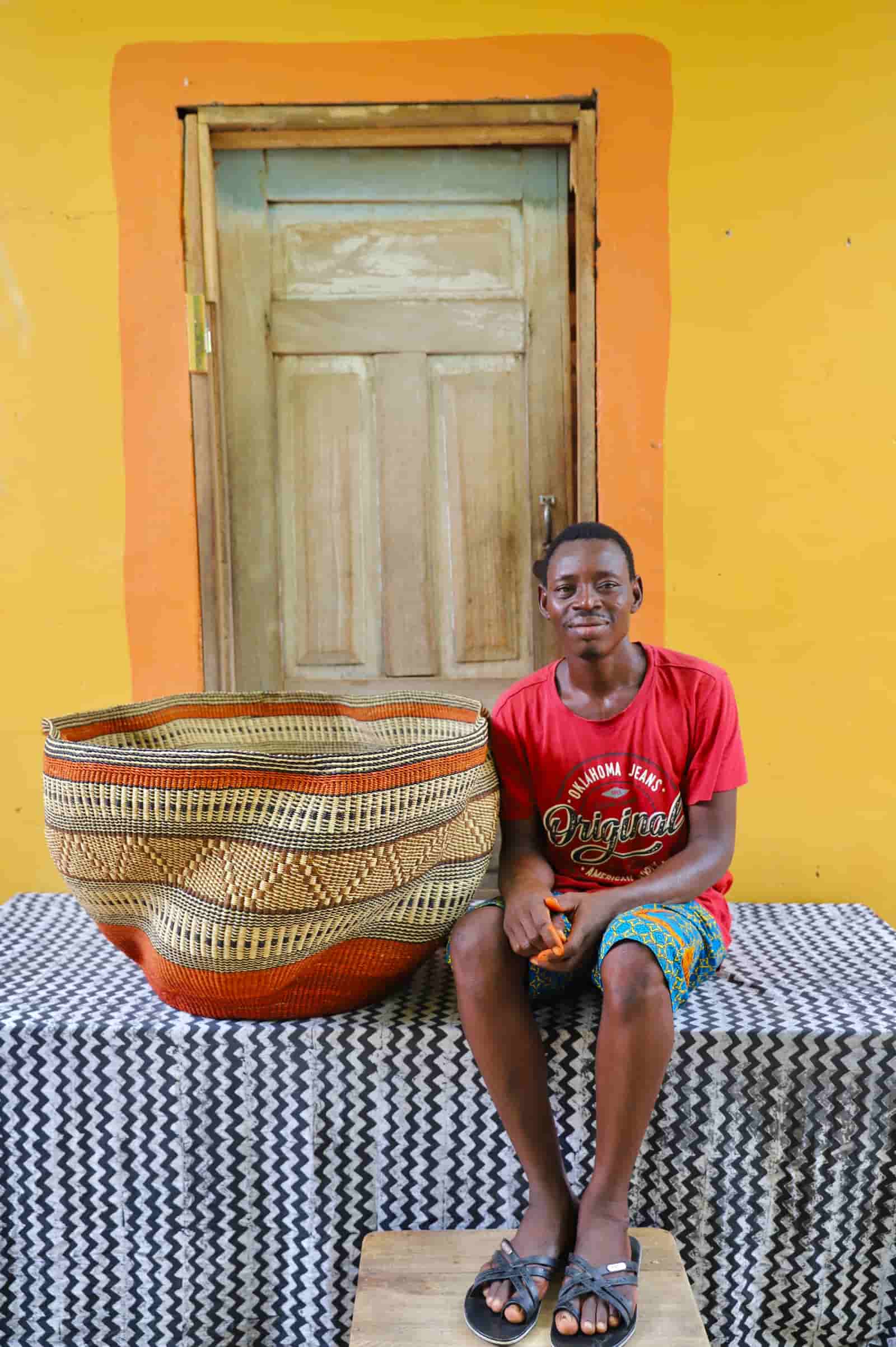 Man sitting on a table with a large woven basket in front of a yellow wall and wooden door.