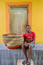 Man sitting with a large Large woven basket with geometric pattern on a black and white checkered tablecloth in front of a wooden door.