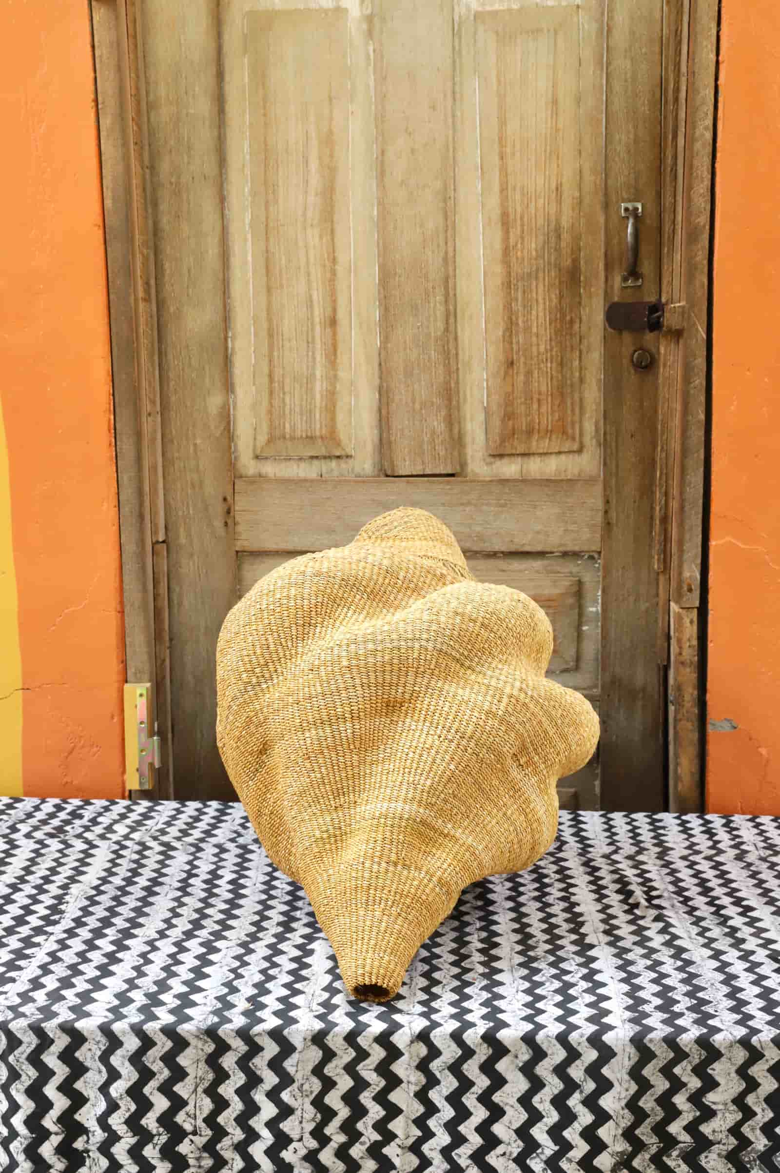 Woven conch lampshade on a black and white patterned tablecloth in front of a wooden door with an orange wall.