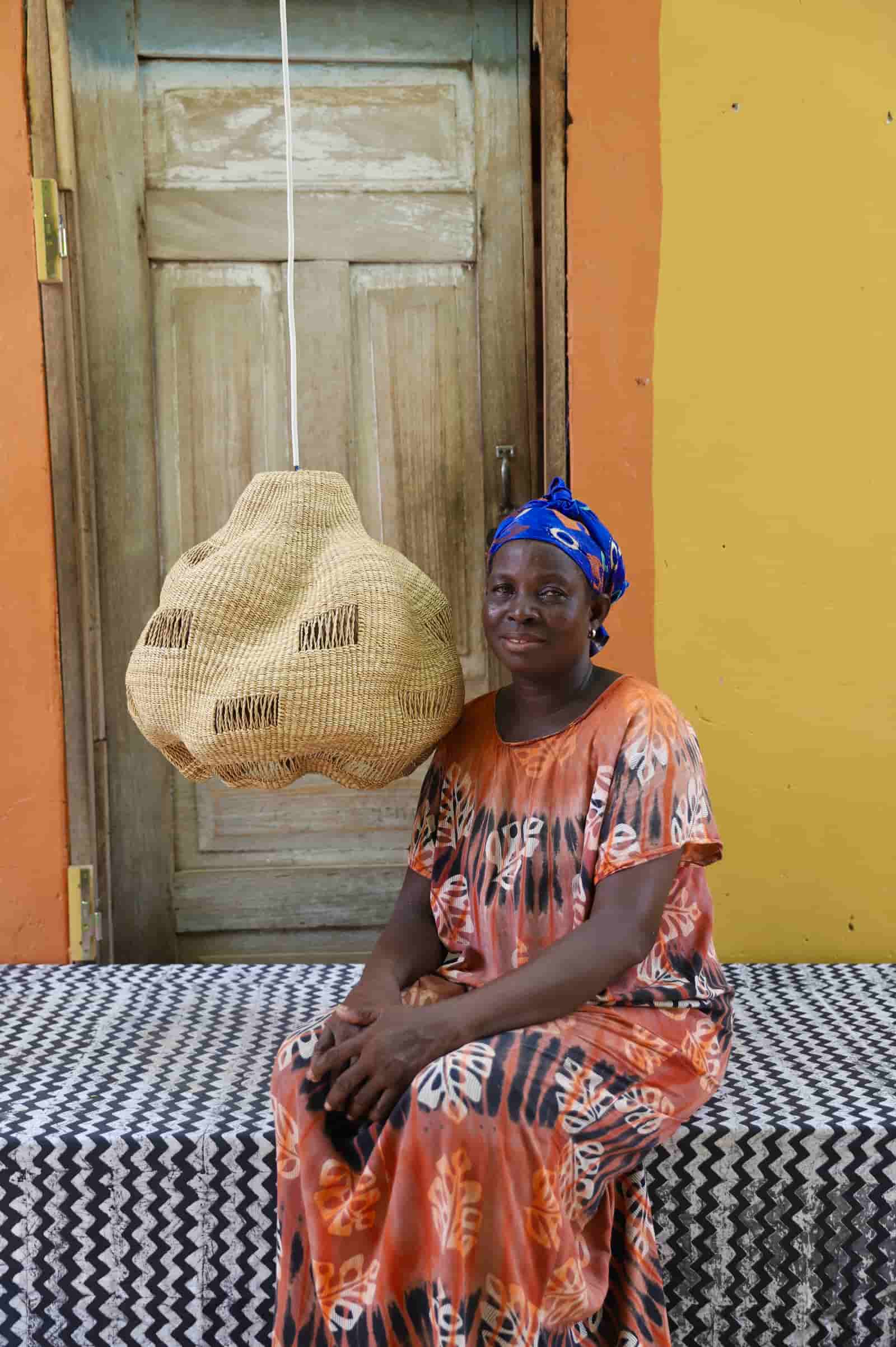 Woman sitting on a bench with a woven basket, wearing an orange dress and blue headscarf, against a colorful wall.