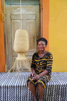 Woman sitting on a bench with a woven lamp behind her against a colorful wall.